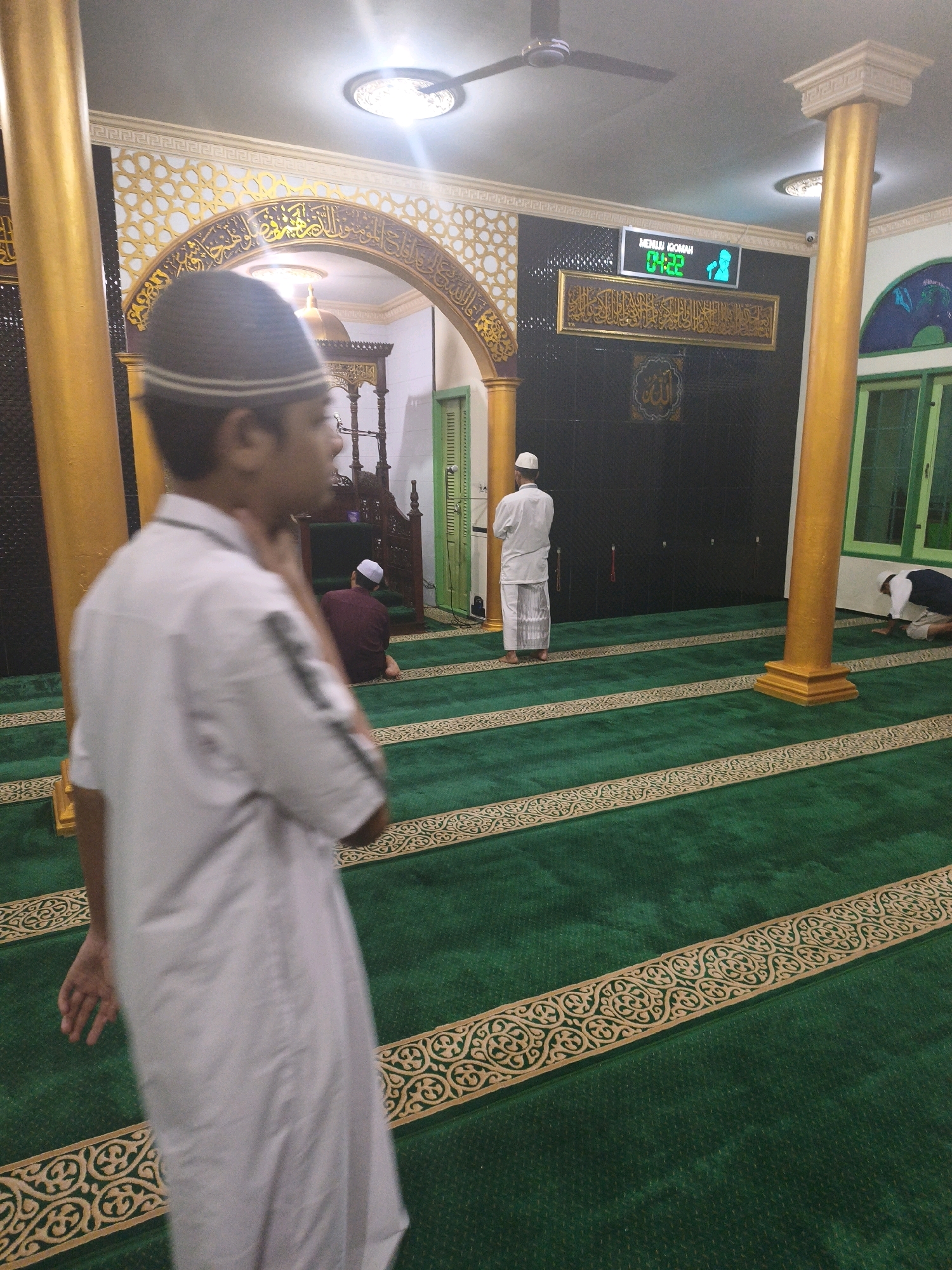 The image depicts the interior of a mosque, likely in Samarinda, Indonesia, judging by the context provided. In the foreground, a young male wearing a white peci (skullcap) and a white peci-like cap is seen from the side, his body blurred as if captured in motion or due to shallow depth of field. He is facing away from the camera, towards the interior of the mosque.
The mosque features a green carpet with decorative golden borders. Several other individuals are present, engaged in prayer or contemplation. One person in a maroon garment is seated on the carpet, facing the qibla (direction of prayer). Further back, two individuals in white are standing. One is facing the qibla, and another is in a prostrating position.
The mosque's architecture includes prominent golden columns and an arched entryway with intricate Arabic calligraphy and geometric patterns. Above the entryway, a digital clock displays "MENUJU IQOMAH" (Approaching Iqomah) and the time "04:22". A large golden calligraphy plaque is mounted on the black wall behind the praying individuals. The lighting appears to be from overhead fixtures and possibly natural light from windows, creating a warm ambiance. The overall scene suggests a moment of religious observance within a well-decorated mosque.
