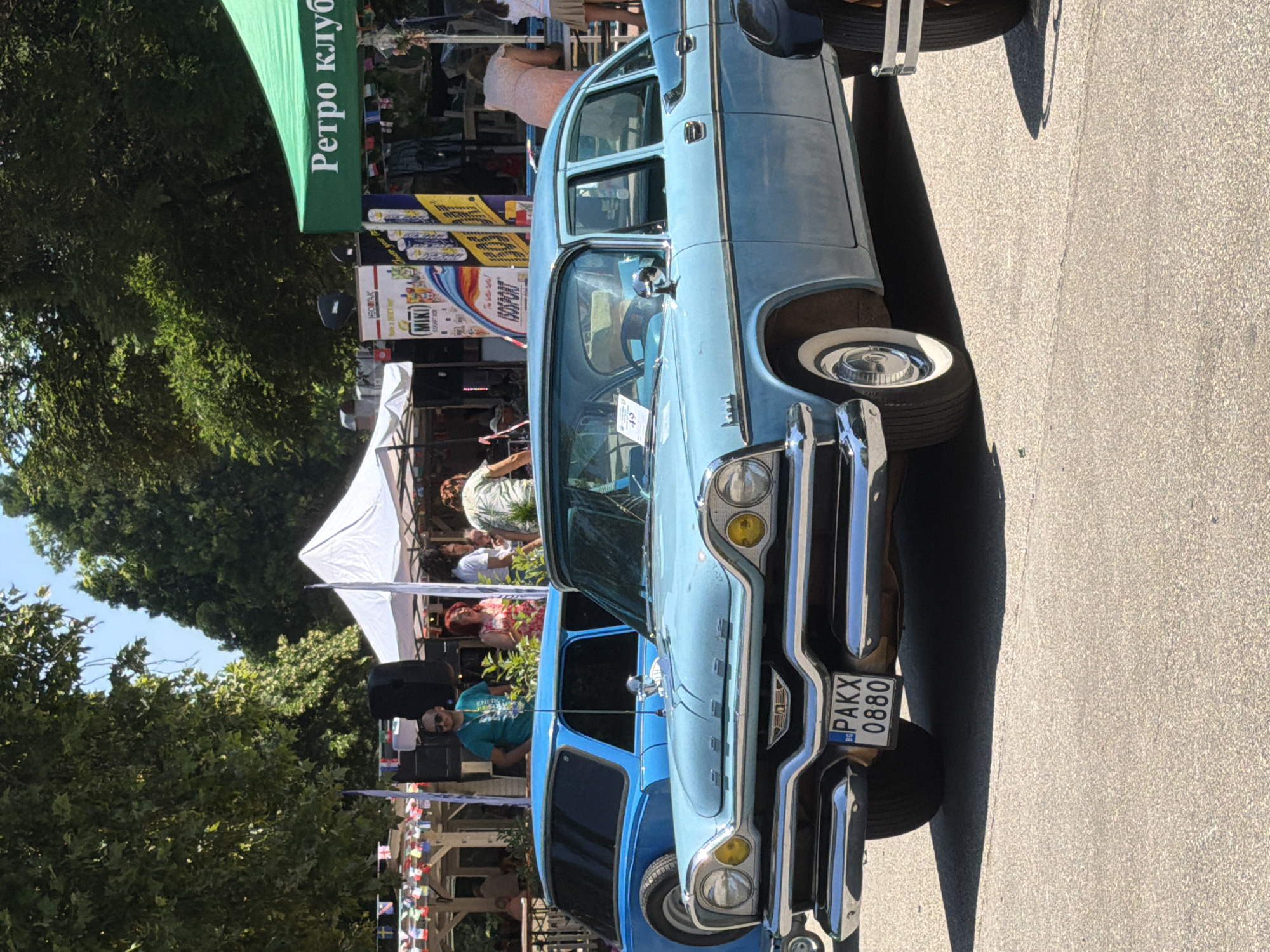 The image shows a vintage light blue sedan parked on an asphalt surface, likely at an outdoor event. In the background, there are trees, tents, and people. A green tent with white lettering "Ретро клуб" is visible on the right. A blue station wagon is parked behind the sedan. The sedan has a license plate reading "BG PAKX 0880". The scene suggests a car show or a festival, possibly in Pazardzhik, Bulgaria given the license plate prefix.