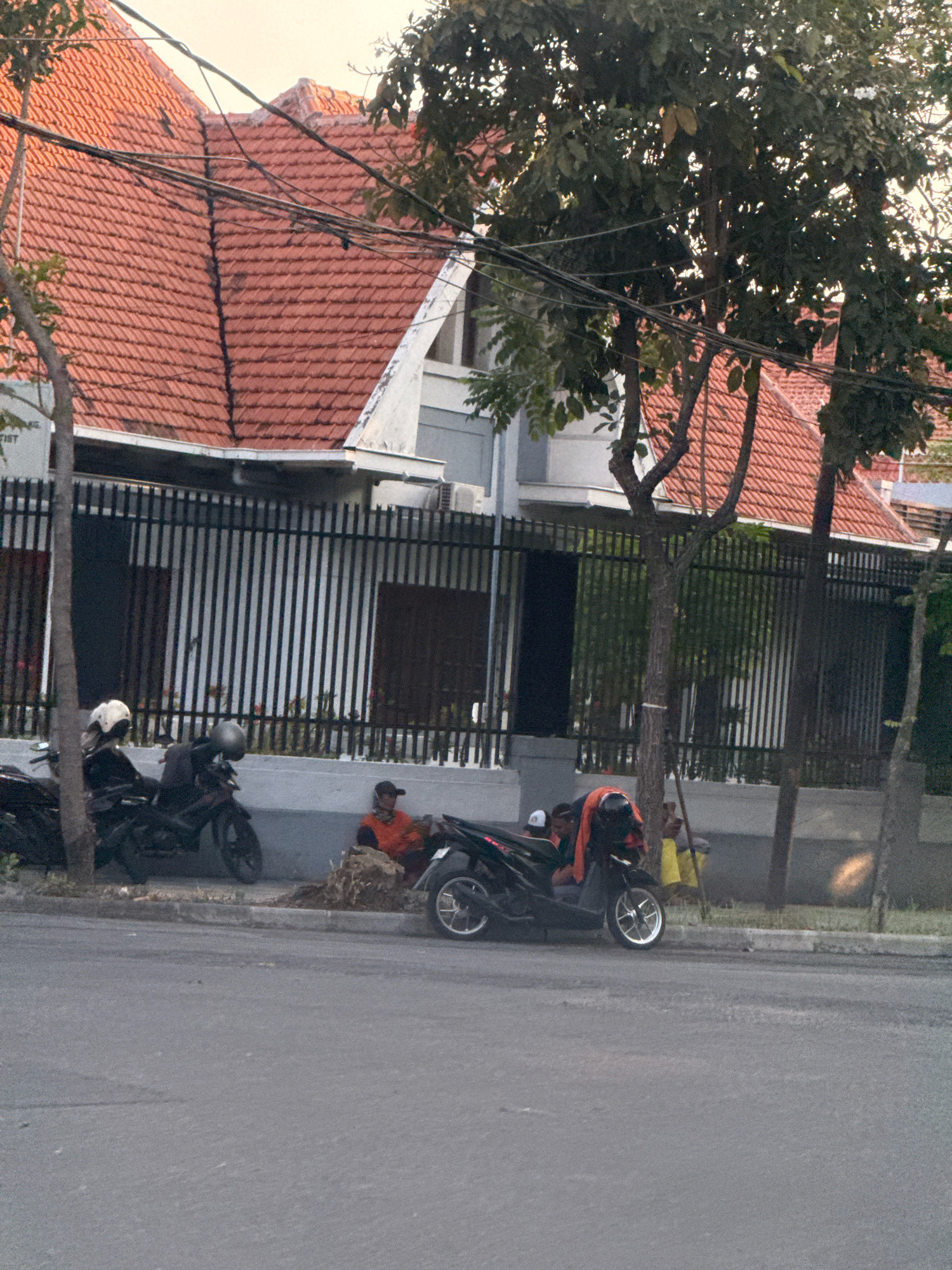 This image depicts a street scene in Surabaya, Indonesia, featuring a residential building with a red tiled roof and a black metal fence. In front of the fence, parked motorcycles are visible, with two individuals seated on one of them. To the right, another person is sitting on the ground next to a pile of debris, wearing an orange shirt and a baseball cap. The street in the foreground is paved and appears to be somewhat worn. Trees with green foliage are present, casting shadows and contributing to the overall atmosphere of the scene. The sky is overcast, suggesting it may be late afternoon or early morning.