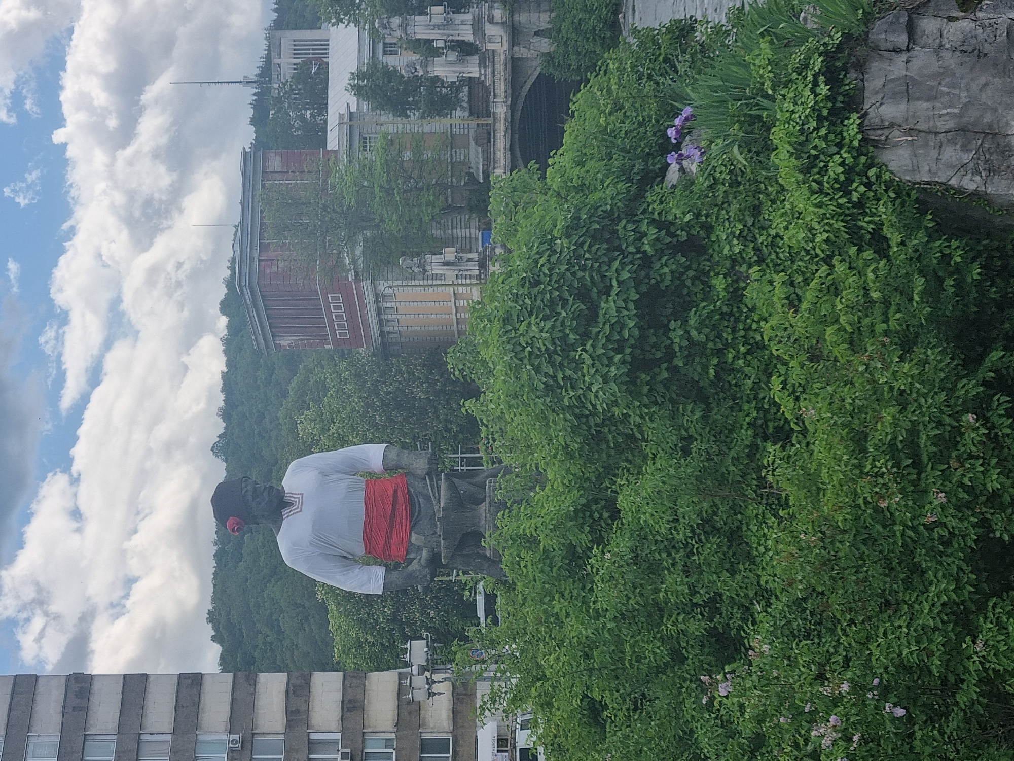 The image shows a statue of Racho Kovacha, the founder of the city of Gabrovo, Bulgaria. The statue is positioned amidst lush green vegetation. In the background, the urban landscape of Gabrovo can be seen, including buildings of various heights and architectural styles. The sky above is partly cloudy.