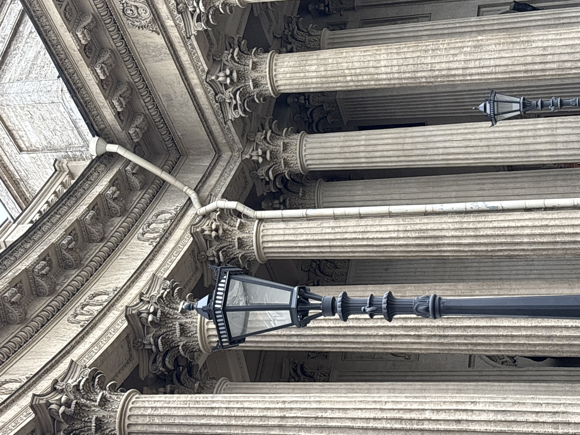 The image shows an architectural detail in Saint Petersburg, Russia. The scene features classical architecture, specifically ornate columns with Corinthian capitals. The building is light-colored, possibly marble or stone. A black street lamp with a multi-sided glass enclosure is mounted on one of the columns. A white pipe runs along the building's surface. The camera angle is from below, looking up at an angle. The sky is not visible.