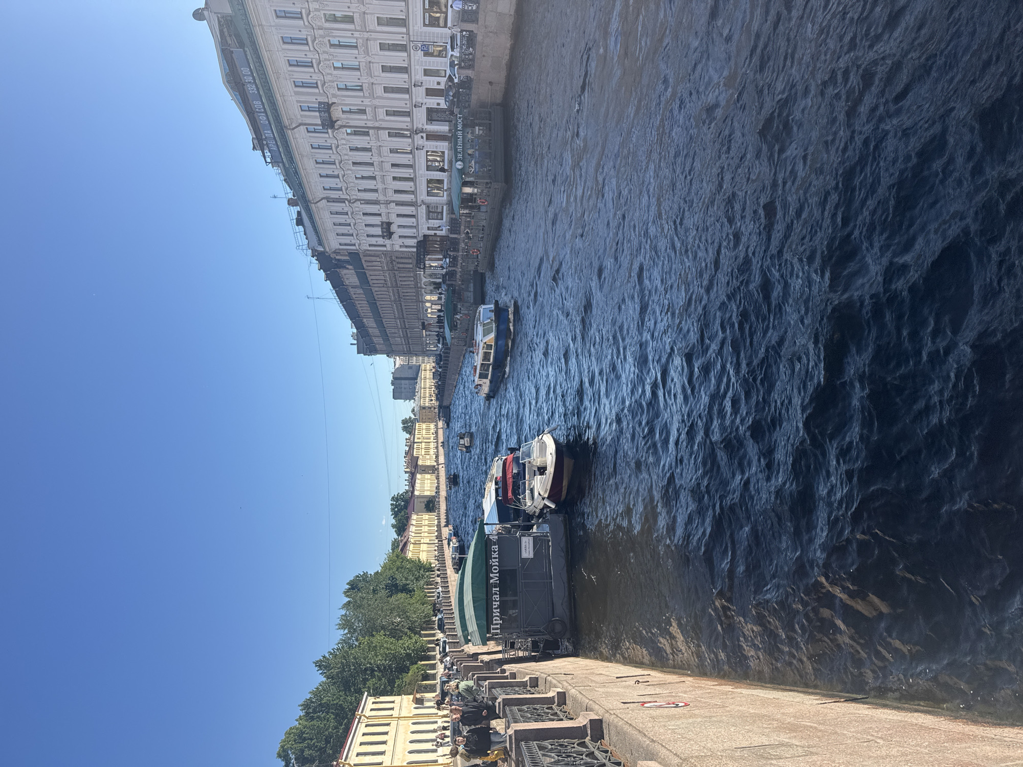 The image shows a view of the Moika River in Saint Petersburg, Russia, on a clear, sunny day. The river is lined with historic buildings and trees on either side. Several tour boats are visible on the water, with people on board and at the piers. The buildings appear to be a mix of residential and commercial spaces, with ornate architecture typical of the city. The water is a deep blue, with ripples indicating some movement. The sky is a bright, cloudless blue. The overall impression is one of a vibrant, active urban waterway in a city rich with history and culture.