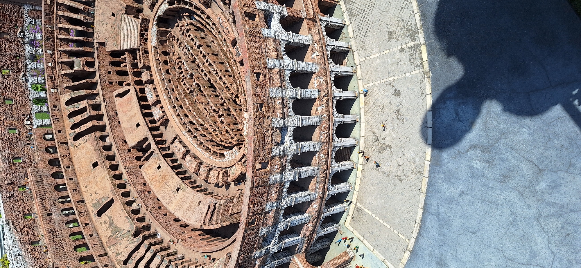 This is a top-down, high-angle photograph of a model of the Colosseum, a famous Roman amphitheater. The model is detailed, showing the tiered seating, arches, and interior structure of the ancient building. The exterior is constructed of brick and stone, with some white stone detailing on the lower levels. The ground surrounding the model is paved with cobblestones, and a darker asphalt-like material is visible in the foreground, casting a large shadow of a person onto it. Scattered across the cobblestone area are several small, colorful figures representing people, some standing and some appearing to be in motion or interacting. The background shows a blurred view of some greenery and other structures, suggesting this is an outdoor exhibit. The presence of the Colosseum model and the mention of Rimini, Italy, suggest this is likely an exhibit at Italy in Miniature (Italia in Miniatura), a theme park located in Viserbella, a frazione of Rimini, Italy, which features scale models of famous Italian landmarks.