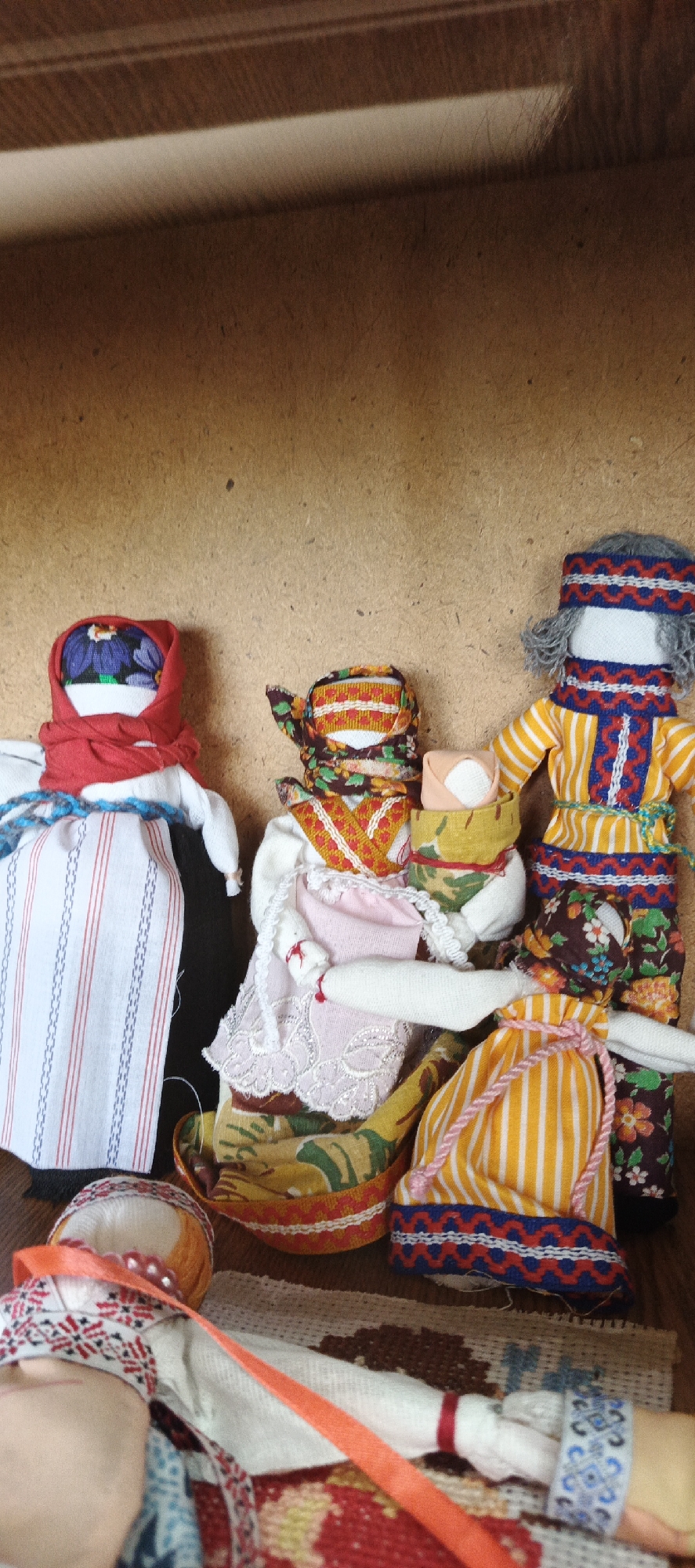 The image features a close-up view of several handcrafted folk dolls, likely Matryoshka dolls or similar traditional Russian dolls, positioned in front of a plain wooden surface. The location is Кемуль, Russia, based on information provided.
The dolls vary in design and attire, reflecting traditional Russian clothing and patterns. One doll wears a red headscarf with floral embellishments, a white dress with red and blue vertical stripes, and a blue crocheted trim. Another doll wears a patterned head wrap with orange and brown hues, an ornate vest with red and orange patterns, and a pale pink underskirt with white lace trim. A third doll is adorned with a grey headwrap with blue and red patterns, a yellow striped shirt with blue ornamentation, and floral print pants.
Additional doll elements include a yellow striped drawstring bag with pink string and a small bundled item. Some dolls rest on a woven rug. The environment is otherwise simple, with the wooden surface providing a neutral backdrop.