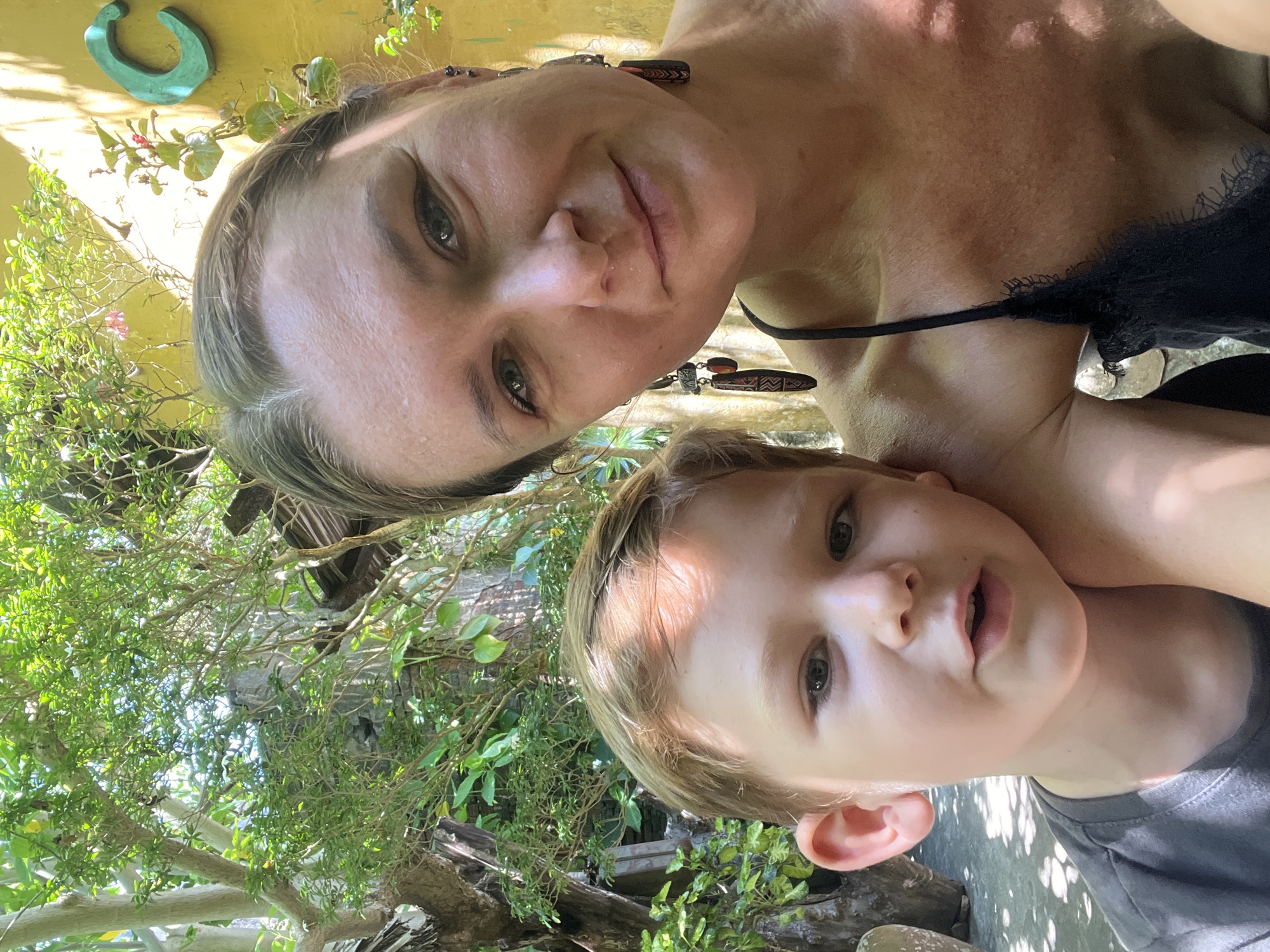 In a medium shot, a woman and a young boy are pictured outdoors, possibly in a courtyard or garden area within Hội An Ward, Vietnam. The woman, on the right, is looking directly at the camera with a slight smile, her face illuminated by sunlight. She has blonde hair pulled back and wears decorative earrings and a black lace-trimmed camisole. The boy, on the left, is closer to the camera and looking slightly away, with his head resting gently on the woman's shoulder. He has light brown hair and is wearing a dark gray t-shirt. Behind them, lush green foliage and trees create a natural backdrop, with dappled sunlight filtering through the leaves. A yellow wall with a large, green decorative letter 'C' is visible in the upper right corner. The ground appears to be paved and shaded by trees.