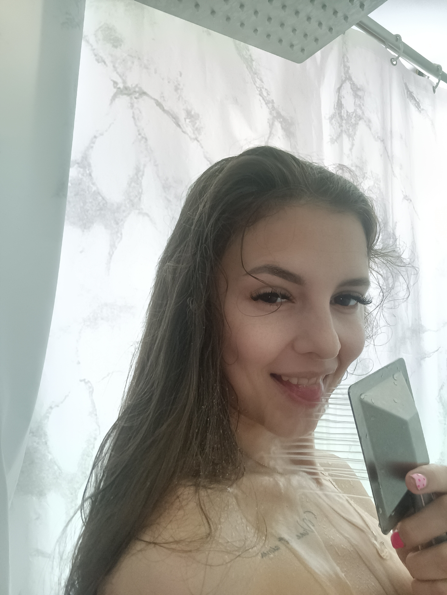 A woman with long brown hair is smiling in a shower, holding a smartphone case. Water is streaming from the shower head and covering her face and chest. The shower curtain is white with a grey marble pattern. The location appears to be a private residence in an unknown city.