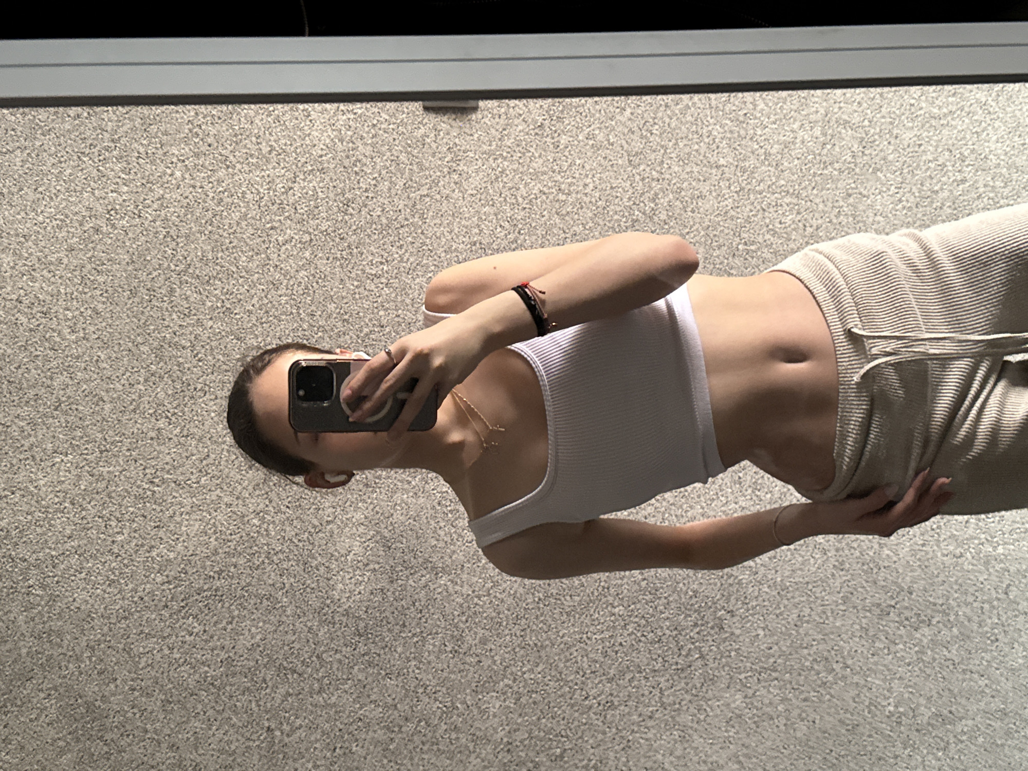 An individual is depicted taking a mirror selfie indoors in Sofia, Bulgaria. The subject's face is obscured by a black smartphone, held by the left hand, which features a prominent camera module and a light-colored case edge. The person is wearing a white ribbed crop tank top and light beige ribbed drawstring pants. A white wireless earbud is present in the visible ear. Two delicate gold-colored necklaces with small pendants adorn the neck. The right wrist displays multiple thin bracelets, one appearing black and red, and a silver-colored ring is worn on the left ring finger. The background consists of a light gray, uniformly textured wall, possibly stucco. A vertical section of a light-colored door or window frame is visible on the right side of the image, reflecting a dark interior space beyond.