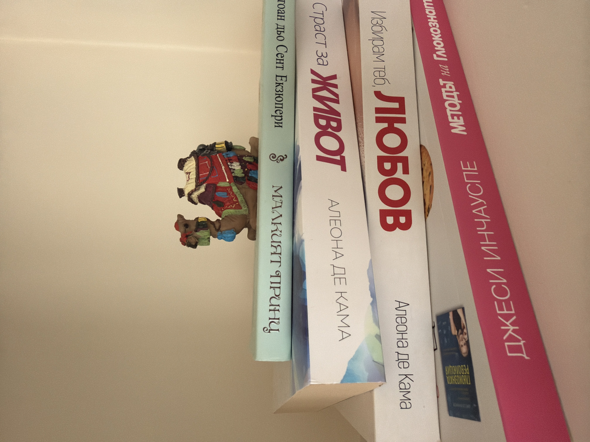 The image shows a stack of books with a small decorative camel figurine on top. The books appear to be in Bulgarian. The bottom book is pink with white text that reads "ДЖЕСИ ИНЧАУСПЕ МЕТОДЪТ на Глюкознат". Above that, a white book has red text that reads "ЛЮБОБОВ' 'Избирам теб.'". The next book up is also white, with red text reading "ЖИВОТ Страст за". The top book is light blue and has white text reading "МАЛАКИТ ПИРПИРЦУ" and other text in Cyrillic script that is partially obscured. The background is a plain, light-colored wall. There is no indication of the location being in Pazardzhik, Bulgaria.
