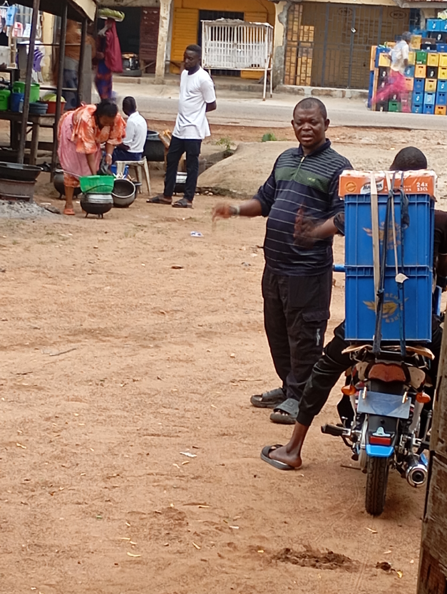 In Shendam, Nigeria, a busy street scene unfolds. A man wearing a striped t-shirt and dark pants stands in the foreground, gesturing with his hands. He is positioned next to a motorcycle equipped with a blue crate carrier laden with orange boxes. Behind him, a woman in a vibrant orange and pink patterned dress is tending to cooking pots, with another person seated nearby. In the background, a man in a white shirt and dark pants stands to the right, observing the scene. Further back, shelves are visible, stocked with various items, and a structure resembling a market stall stands to the left. The ground is covered in a dusty, reddish-brown soil, with scattered debris. Buildings with yellow and brown facades line the street, and stacks of colorful crates are visible in the distance. The overall impression is one of a lively, everyday market environment.