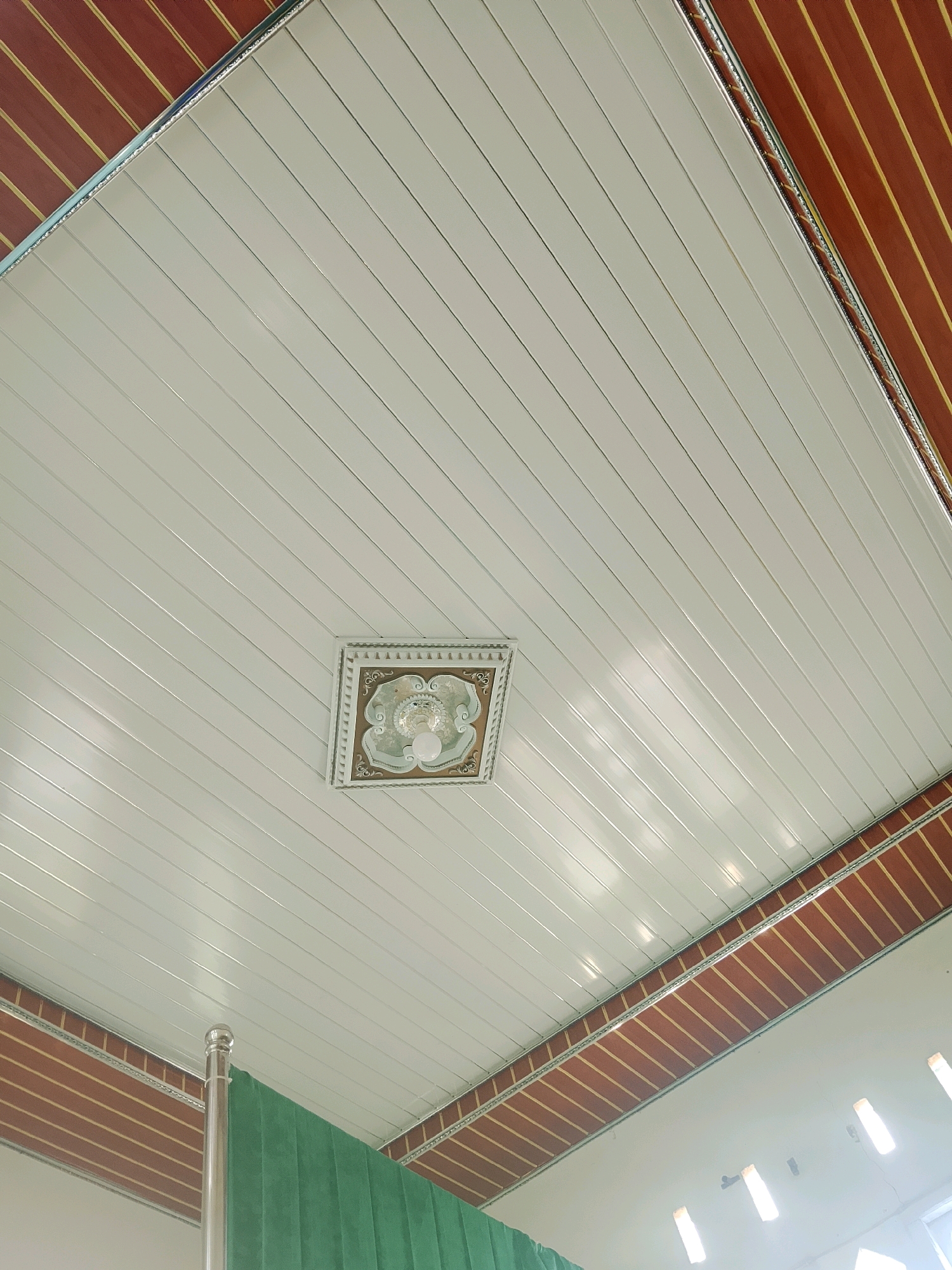 This image depicts a white, panelized ceiling with decorative brown and gold trim along the edges. A square ceiling fixture with intricate patterns and a lightbulb is centrally located. A metallic pole and a section of green fabric are visible in the lower left corner of the image, suggesting an indoor space with some form of partitioning or decorative element. The scene is indoors and does not contain any information about its specific location in Negeri Agung, Indonesia.
