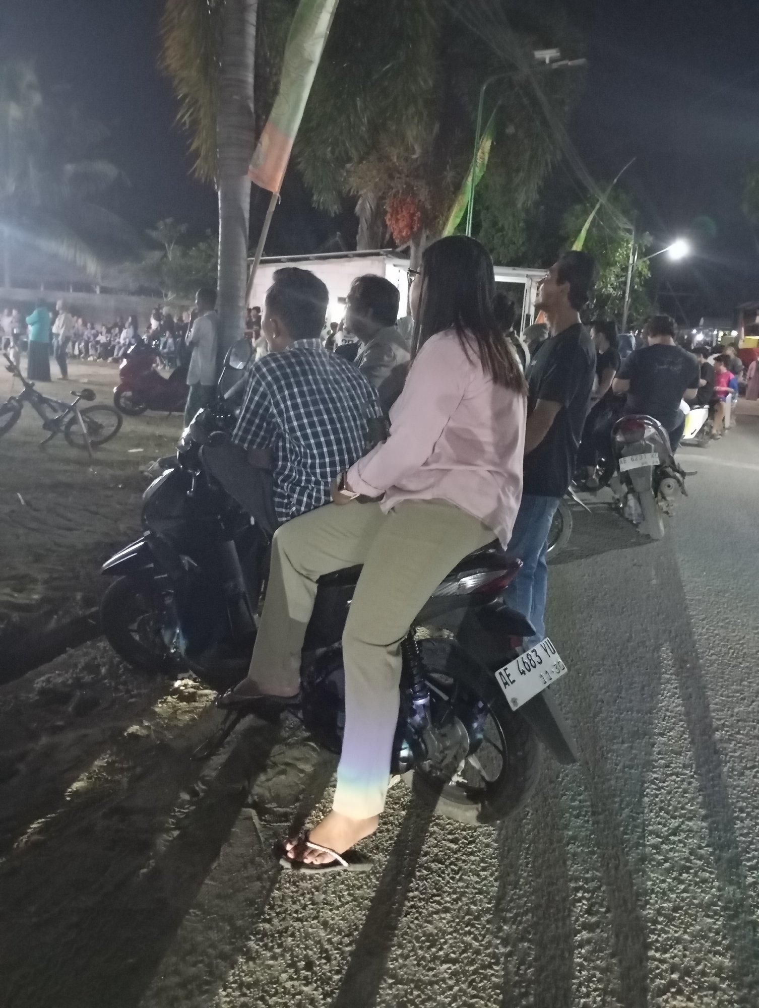 In the city of Pacitan, Indonesia, at night, several individuals are seen gathered. In the foreground, a dark-colored motorcycle is parked on a paved road adjacent to a sandy area. Two people are seated on the motorcycle. The person in the rear, wearing a light pink long-sleeved shirt and beige pants, is facing away from the camera. The person in front, wearing a blue and white checkered long-sleeved shirt, is also facing away. Behind them, more people are visible, some standing and some on other motorcycles. In the background, palm trees and other foliage are illuminated by artificial lights. The scene appears to be a public gathering or event.