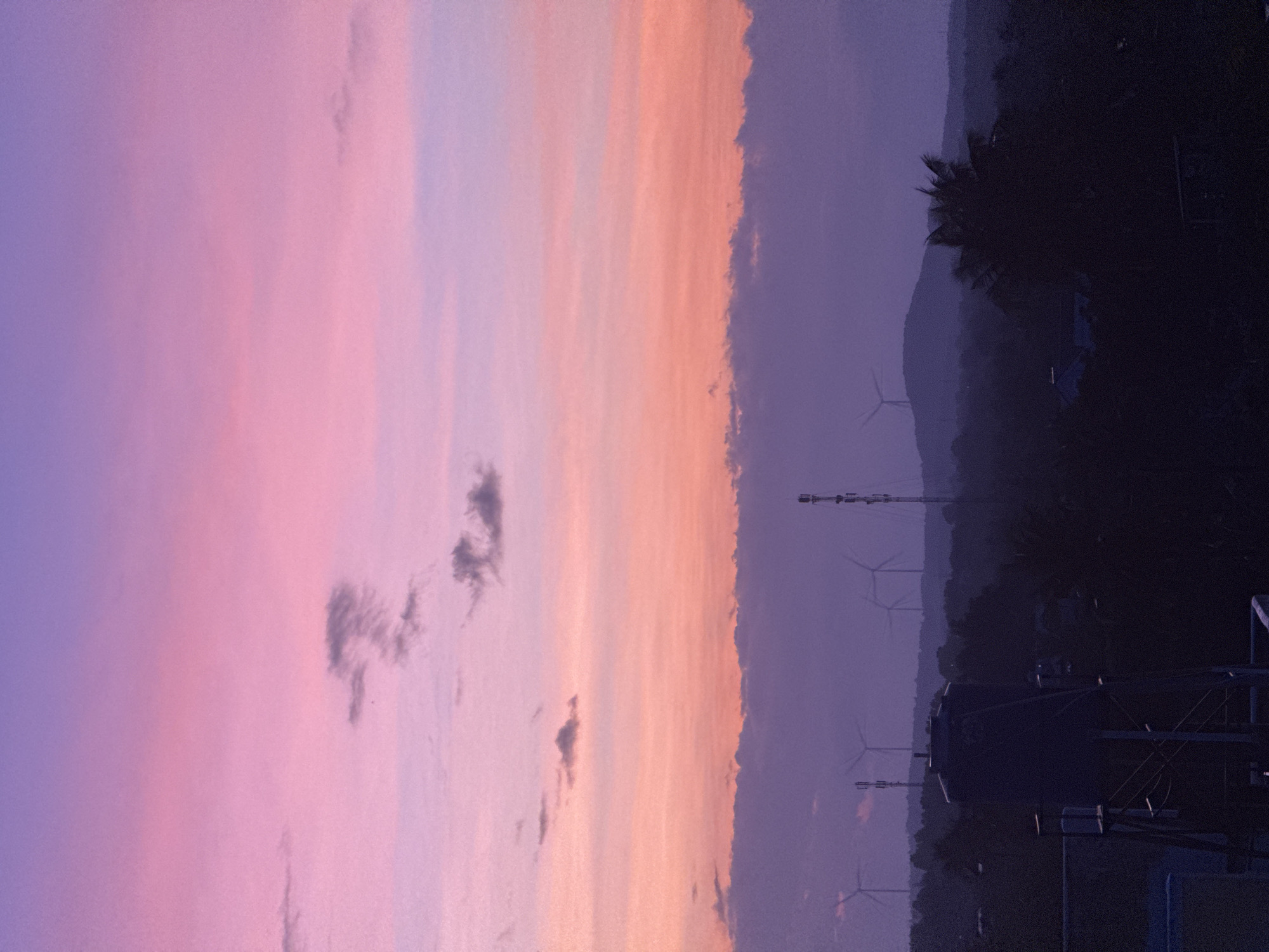 The image captures a dusky sky over Mũi Né, Vietnam. The sky is a gradient of soft purple and pink, with wisps of clouds scattered across it. In the foreground, silhouetted against the vibrant sky, is a dark landscape of trees and hills. Several wind turbines are visible on the horizon, their blades blurring in the dim light, suggesting the presence of a wind farm. A tall communication tower stands out amongst the turbines. In the extreme foreground, the top of a dark water tank and some scaffolding are visible, indicating a man-made structure in an elevated position. The overall atmosphere is serene and slightly ethereal, characteristic of a sunset or twilight in a coastal area.