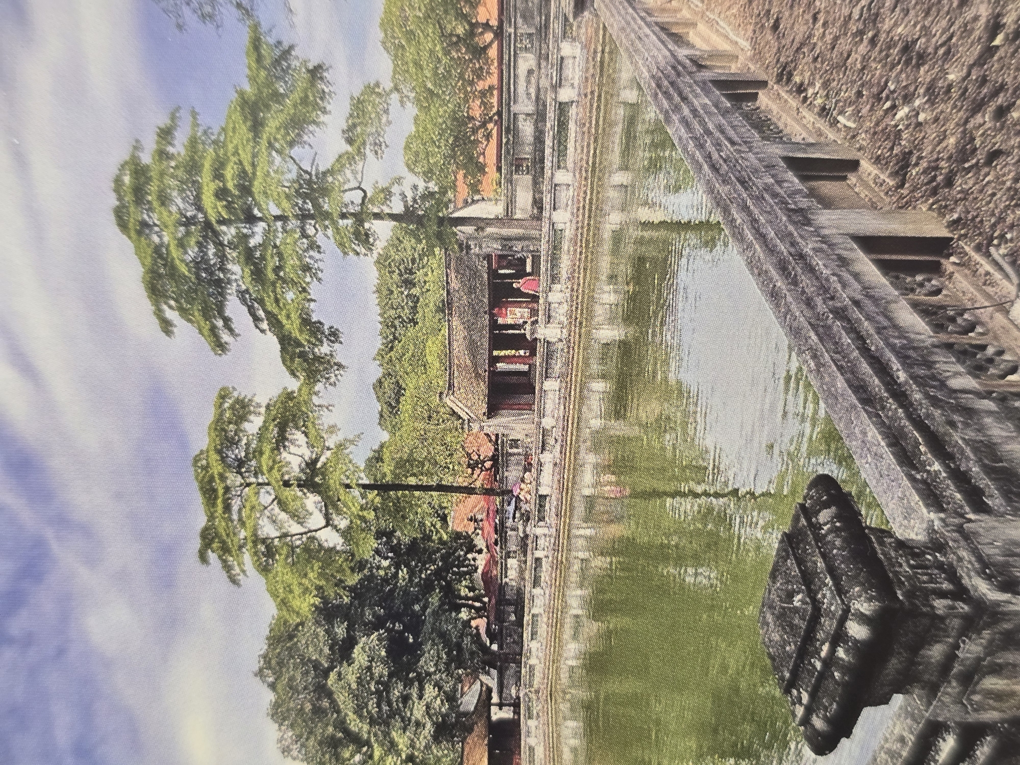 The image presents a tranquil outdoor scene featuring a large, rectangular body of water, likely a pond or canal, that occupies the foreground and middle ground. On the right side, a series of textured stone steps or a terraced pathway with low walls descends towards the water. A dark, carved stone block or pedestal is partially visible in the immediate foreground, positioned at the water's edge.
Across the calm, greenish water, a traditional East Asian-style building is prominently featured. It possesses a distinctive reddish-brown tiled roof and light-colored walls, indicative of historical architecture such as a temple or pagoda. The building is surrounded by an abundance of lush, green trees and dense foliage. Two particularly tall trees with slender trunks and long, drooping branches stand out on the left side, their forms creating elegant reflections on the water's surface.
A few small figures, possibly people, can be discerned near the building, suggesting visitors or inhabitants. One figure, partially visible, appears to be wearing a red garment. The sky above is bright blue with scattered white clouds, indicating a clear day, likely late morning or early afternoon.
The architectural style and vegetation strongly suggest an East Asian setting, such as Vietnam. This visual evidence contradicts the provided location context of Sofia, Bulgaria. No discernible text is visible in the image.