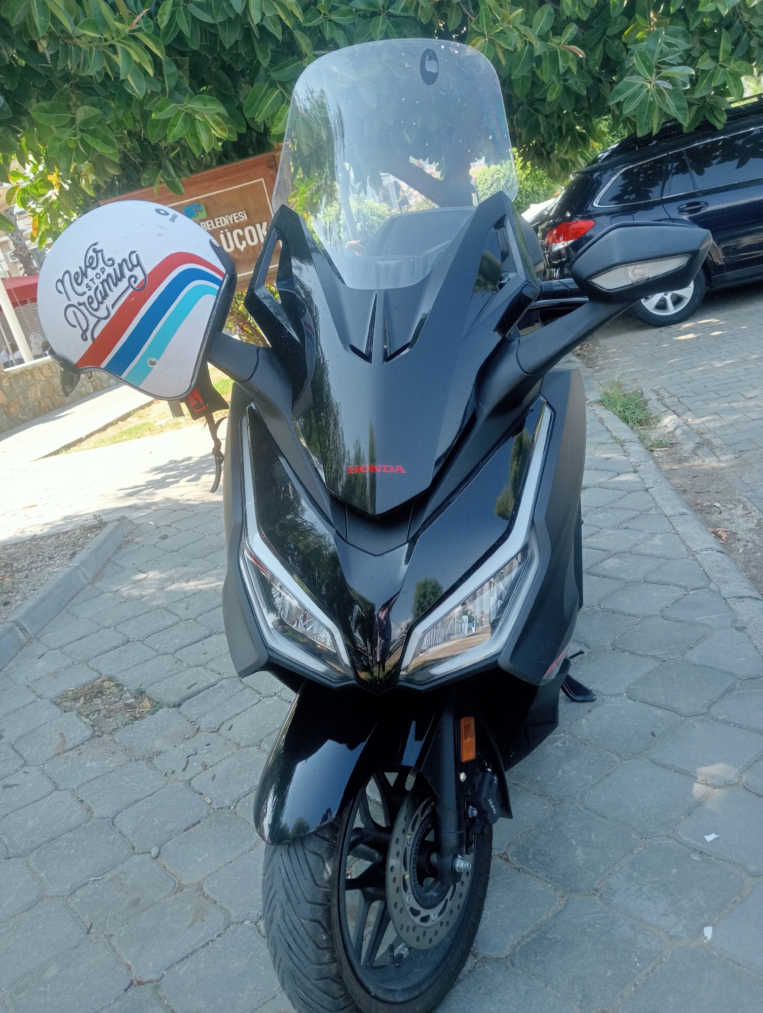 The image shows a black Honda scooter parked on a cobblestone street in Fethiye, Turkey. A white helmet with red, white, and blue stripes and the text "Never Stop Dreaming" is resting on the handlebars. In the background, a dark-colored SUV is parked behind some green foliage, and a sign with Turkish text that reads "BELEDİYESİ UÇOK" is visible. The lighting suggests it is daytime with bright sunlight.