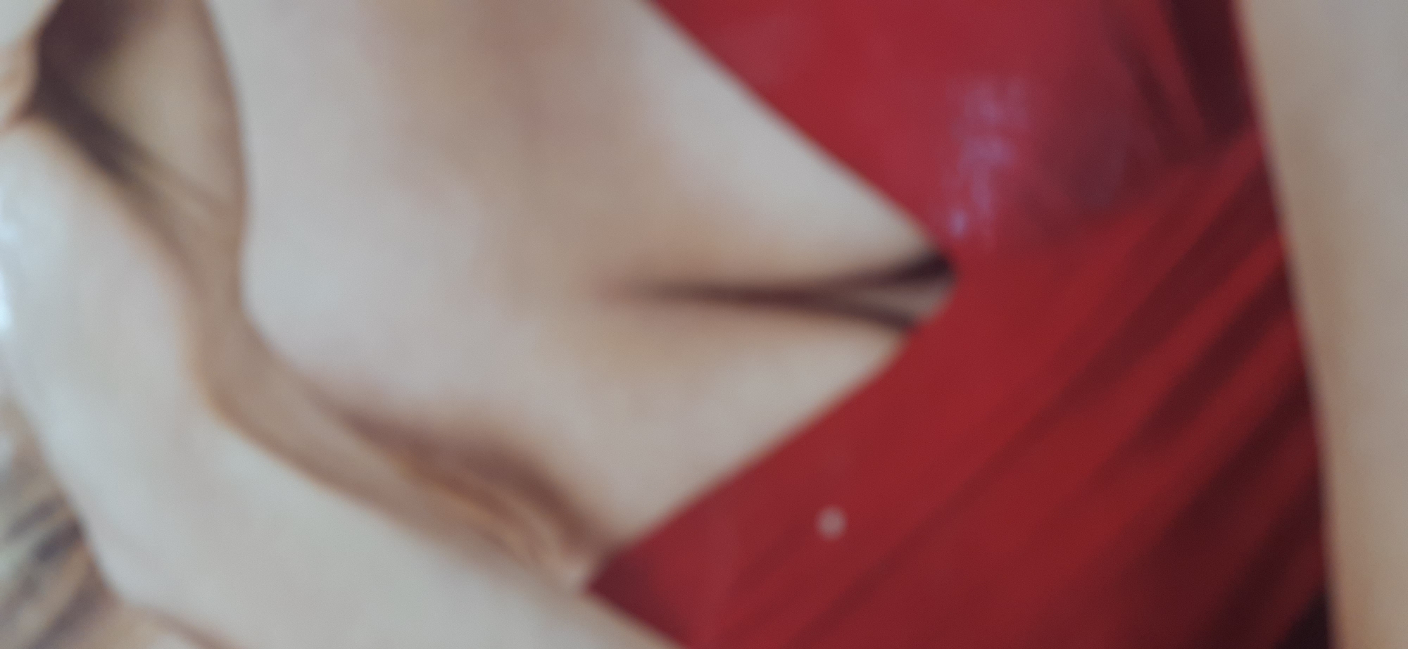 The image is a close-up, blurred, and angled shot of a person's chest and upper torso, wearing a red garment that appears to be a dress or top. The garment has a V-neckline that reveals cleavage. There is a faint, small white circular embellishment visible on the left side of the red garment. The lighting is soft, and the background is out of focus, but suggests a light-colored surface. There are no identifiable landmarks or contextual clues to indicate a specific location within Dnipro, Ukraine, or any other city.