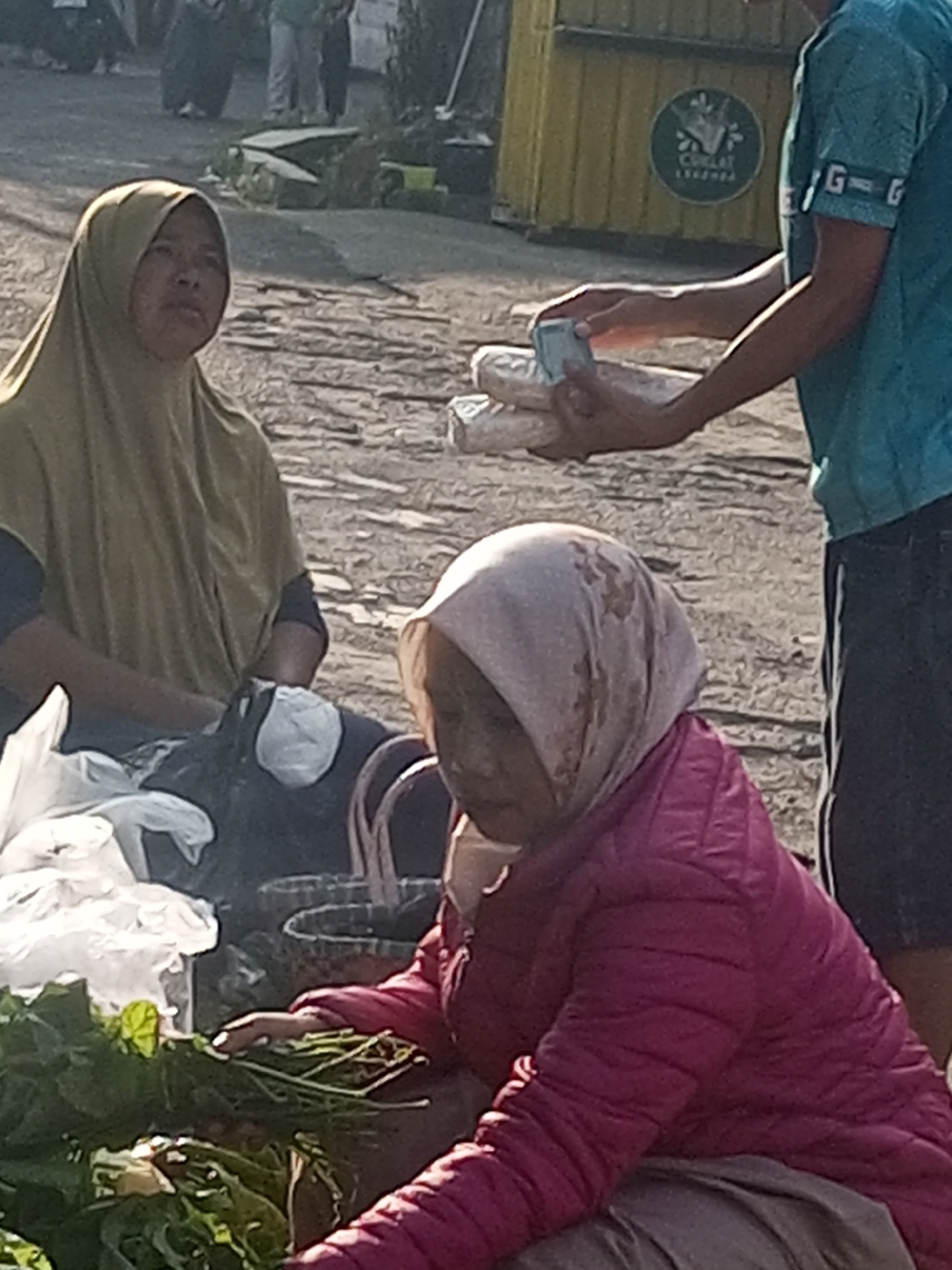 In Krasak, Indonesia, two women are seen at what appears to be a market stall. The woman on the left wears a light brown hijab and a dark top. The woman on the right wears a light pink hijab and a vibrant pink puffer jacket. She is leaning forward, tending to a pile of green leafy vegetables and long green stalks in front of her, possibly preparing them for sale. Behind the woman on the right, another person, partially visible and wearing a blue t-shirt and dark shorts, is holding a stack of white, cylindrical objects, possibly food items like rice cakes or bread, and appears to be engaged in a transaction. A yellow food stall with a circular green logo is visible in the background. The ground is paved with rough stones, and the overall environment suggests an outdoor market setting.