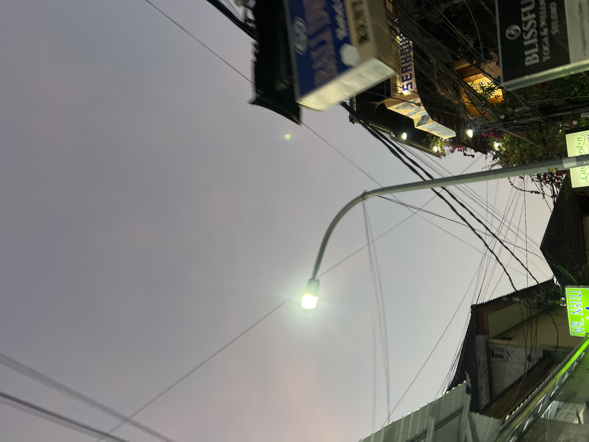 The image captures a street scene in Kuta, Indonesia, during twilight. A street lamp with a bright light illuminates the dusky sky, which is a soft gradient of purple and grey. Overhead, a complex network of electrical wires crisscrosses the frame, indicating a dense urban environment. On the right side of the image, a multi-story building with a dark, sloped roof is visible. A prominent blue sign with white lettering, partially obscured, reads "BALI D..." suggesting it might be for a business. Below this, another sign reads "SEAH..." further identifying a commercial establishment. The building is adorned with lights and flowering vines with red blossoms, adding a touch of color to the scene. On the far left, another building with a sloped roof is partially visible, along with a green sign that says "WR. NAULI". In the foreground, parts of a metal awning and more electrical wires are present. The overall impression is of a bustling commercial street in Kuta at dusk, characterized by its dense infrastructure and vibrant signs.
