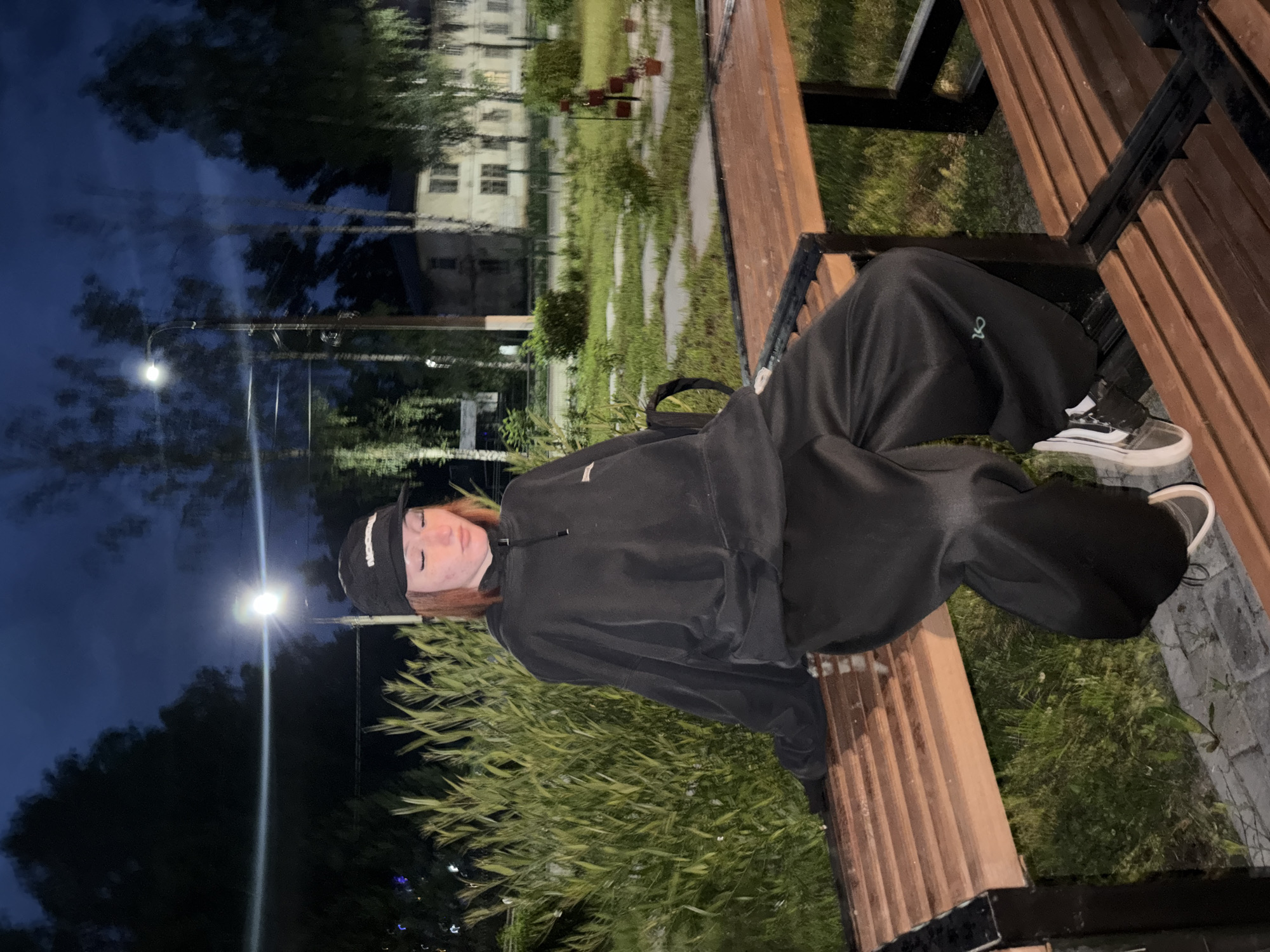 A young female with auburn hair, wearing a black baseball cap, black hoodie, and black sweatpants with a logo on the left thigh, sits on a wooden park bench at night. She is positioned in the foreground, with her eyes closed and head tilted down. A street lamp illuminates the scene, casting a bright glow. Behind her, a dense green hedge borders the grassy area. In the background, trees with visible branches and leaves are sparsely scattered, and a light-colored, multi-story building is visible. The environment appears to be a residential area within the city of Chaikovsky, Russia, possibly a courtyard or park.