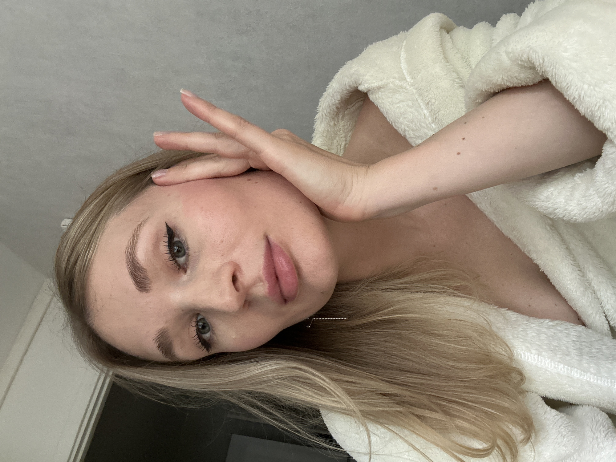 This is a close-up, vertically oriented, eye-level, indoor photograph of a young woman. She has long, blonde hair, blue eyes, and fair skin. She is wearing a white, fuzzy bathrobe, and her hand is resting on her forehead. Her makeup includes eyeliner and mascara. The background is a grey wall. The image appears to be taken during the daytime, possibly in a home setting. No text is visible.