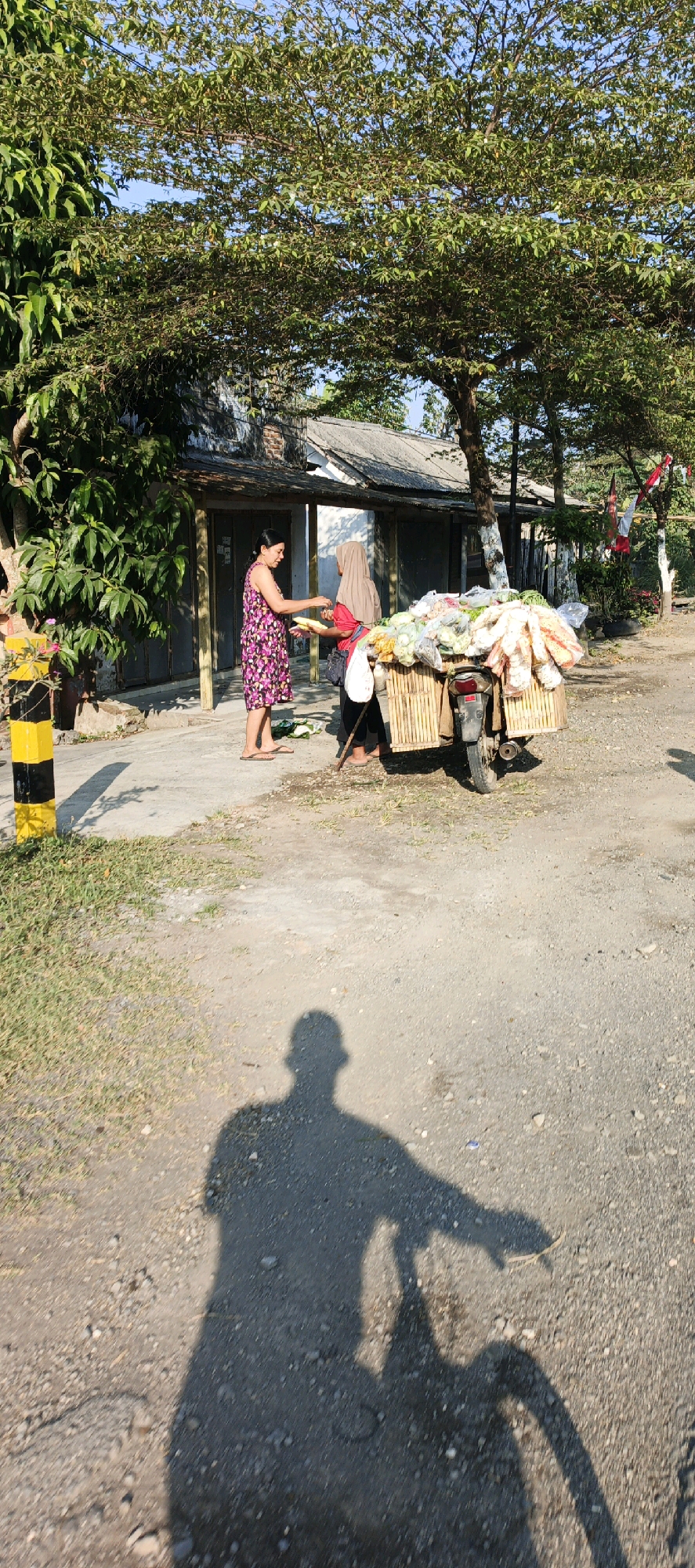 A woman in a floral dress is buying vegetables from a vendor in Mlokorejo, Indonesia. The vendor, wearing a hijab, is standing behind a motorcycle loaded with produce. The scene takes place on a dusty road in front of a rustic building. A shadow of a person on a bicycle is visible in the foreground.