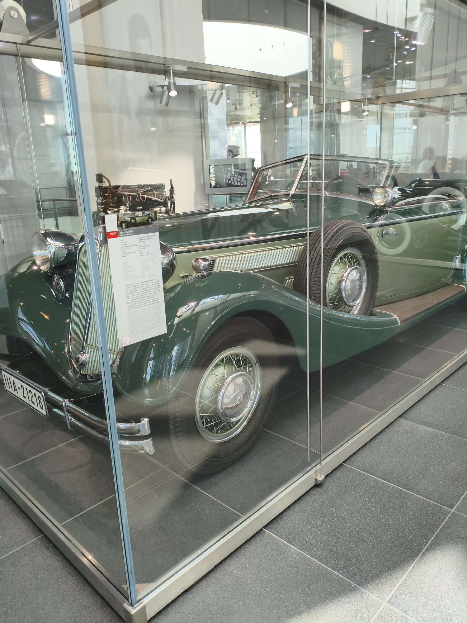 The image shows a vintage green Horch 853 Cabriolet automobile from 1937, displayed inside a glass enclosure in the Audi Forum Ingolstadt in Ingolstadt, Germany. The car is a convertible with a spare tire mounted on the side. A white informational plaque is attached to the front grille of the car. The enclosure is made of glass panels supported by metal framing. The floor is tiled with dark gray tiles. The background is blurred, showing a museum-like environment with lighting fixtures and other exhibits. No people are clearly visible in the immediate vicinity of the car.