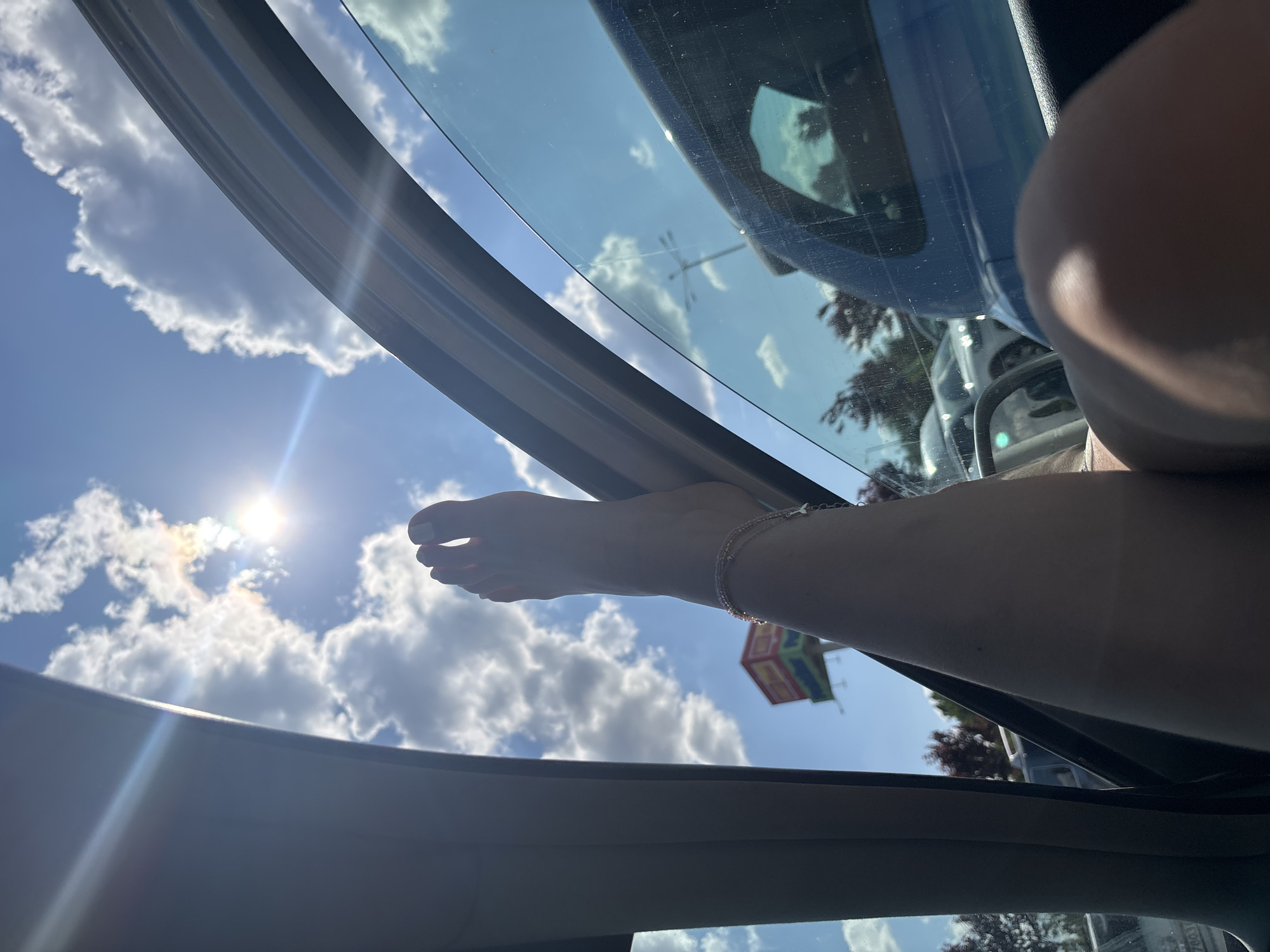 The image is captured from inside a vehicle, looking outwards through an open side window. A bare human leg and foot, displaying white-painted toenails and a multi-strand anklet above the ankle, is propped on the lower frame of the open window. The bright blue sky, filled with white cumulus clouds, dominates the upper exterior view. The sun is visible, creating a strong lens flare across the upper left portion of the image.
In the mid-ground, beyond the vehicle, other cars are parked in what appears to be a commercial parking lot. Dark-leafed trees are visible, along with a utility pole and overhead wires. A prominent, tall, multi-colored sign or billboard structure stands in the distance. The sign features distinct red sections at the top and bottom, separated by a green and yellow central area. Partially visible text on the green section of this sign reads "MR.Bricolage," confirming the location as a MR.Bricolage store in Sofia, Bulgaria. The interior of the vehicle, including parts of the dashboard and door panel, is visible in the foreground.