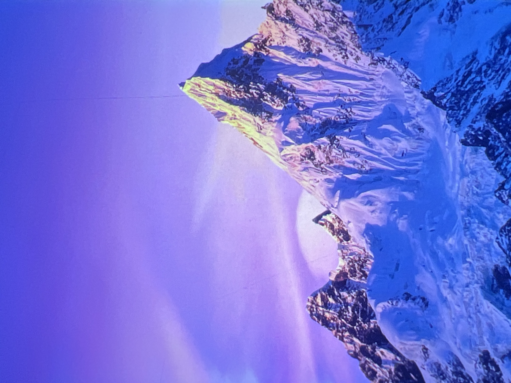 The image shows a majestic snow-capped mountain range against a vibrant purple sky. The peaks are sharp and jagged, with the tallest mountain in the foreground catching the sunlight on its summit, creating a golden glow. The snow on the mountains is a mix of white and deep blue hues, suggesting shadows and the play of light. Wispy clouds are visible in the sky, adding texture to the purple backdrop. The overall scene evokes a sense of grandeur and natural beauty. There are no people, specific landmarks, or urban elements visible in the image.