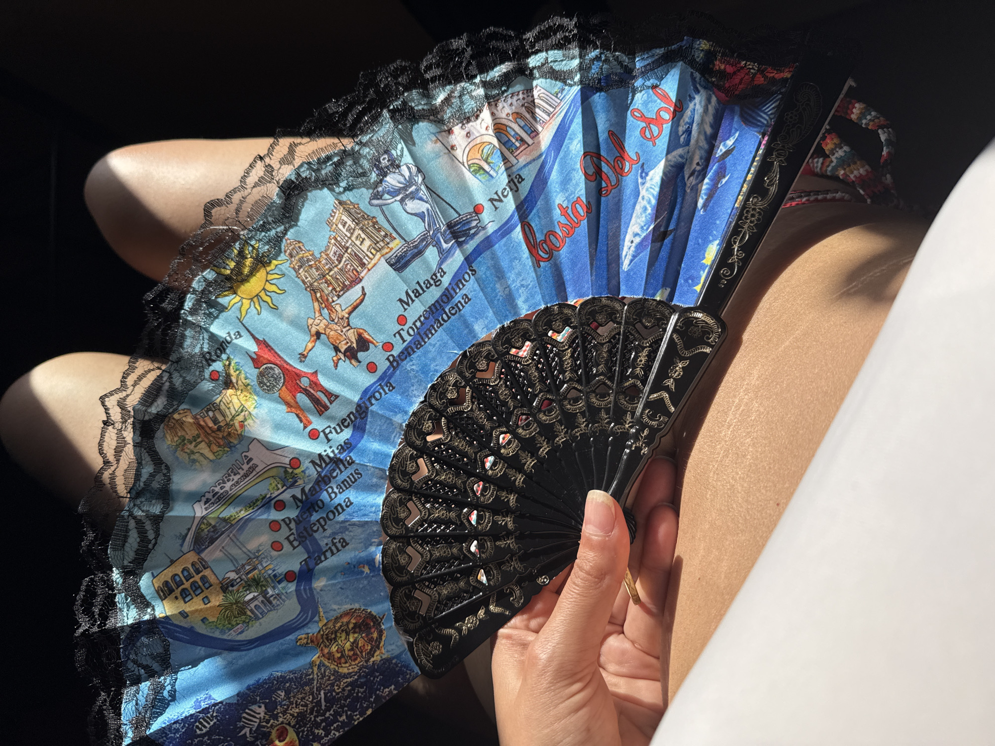 A person is holding a souvenir fan in their hand, in an indoor setting. The fan is partially open, displaying the text "Costa Del Sol" and names of cities and regions like "Ronda," "Malaga," "Torremolinos," "Fuengirola," "Marbella," and "Tarifa" printed on a blue background with illustrative images. The hand holding the fan has a gold ring on one finger. The scene is lit by sunlight, creating highlights and shadows on the hand, fan, and background.