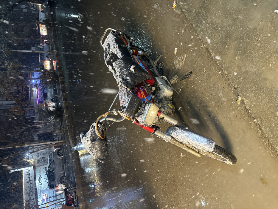 The image depicts an urban street scene at night during a snowfall, with a motorcycle lying on its side in the foreground. The red and black motorcycle is heavily coated with white snow or sleet, particularly on its seat, engine cover, handlebars, and front fender, indicating active and accumulating precipitation. The ground is wet and dark, reflecting the artificial lights. Snowflakes are visibly falling across the entire frame.
In the mid-ground and background on the left, several buildings line the street, displaying illuminated windows and signage. Parked cars are visible, their lights creating reflections on the glistening road surface. Bare trees stand along the roadside, possibly also dusted with snow.
Visible text on a prominent banner includes the English words "Zaiko Waters." Below and around this, there is text in Urdu or Persian script. One word clearly discernible beneath "Zaiko Waters" appears to be "پانی" (Pani), meaning water, consistent with the English text. Another banner shows what looks like "دانہ" (Daana) or similar script, likely part of a business name. The scene conveys a cold, wintry atmosphere in an urban setting, consistent with Quetta City Tehsil, Pakistan. The fallen motorcycle is the central subject, suggesting an incident or simply being laid down in the snowy conditions.