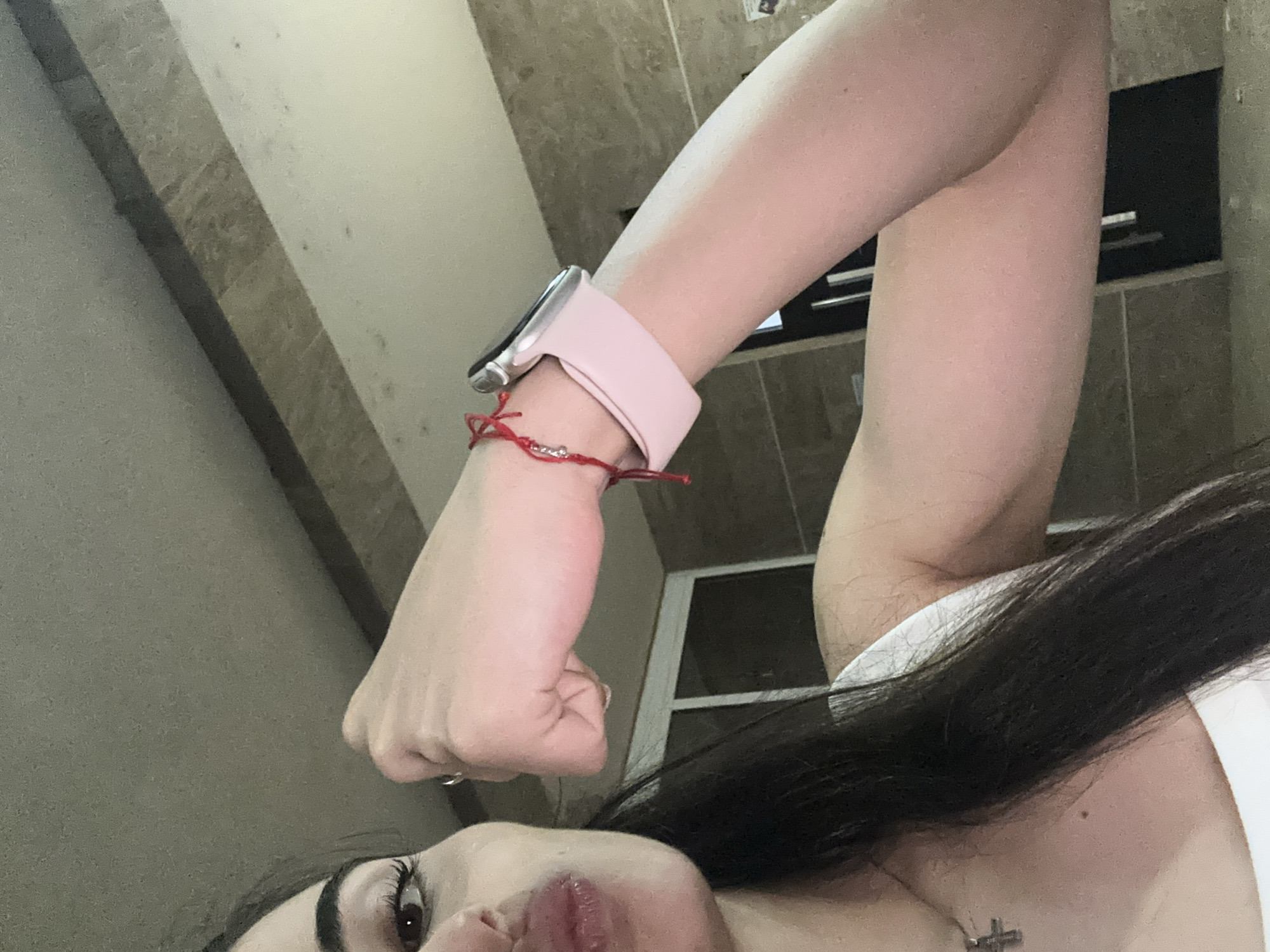 A close-up shot shows a woman's right arm and hand. She is wearing a pink smartwatch with a red string bracelet on her wrist. Her fist is clenched, and her arm is flexed. In the foreground, a portion of her face is visible, with her lips pursed and her left eye looking towards the camera. Her dark hair falls over her shoulder. The background appears to be an indoor setting with tiled walls and a dark doorway.