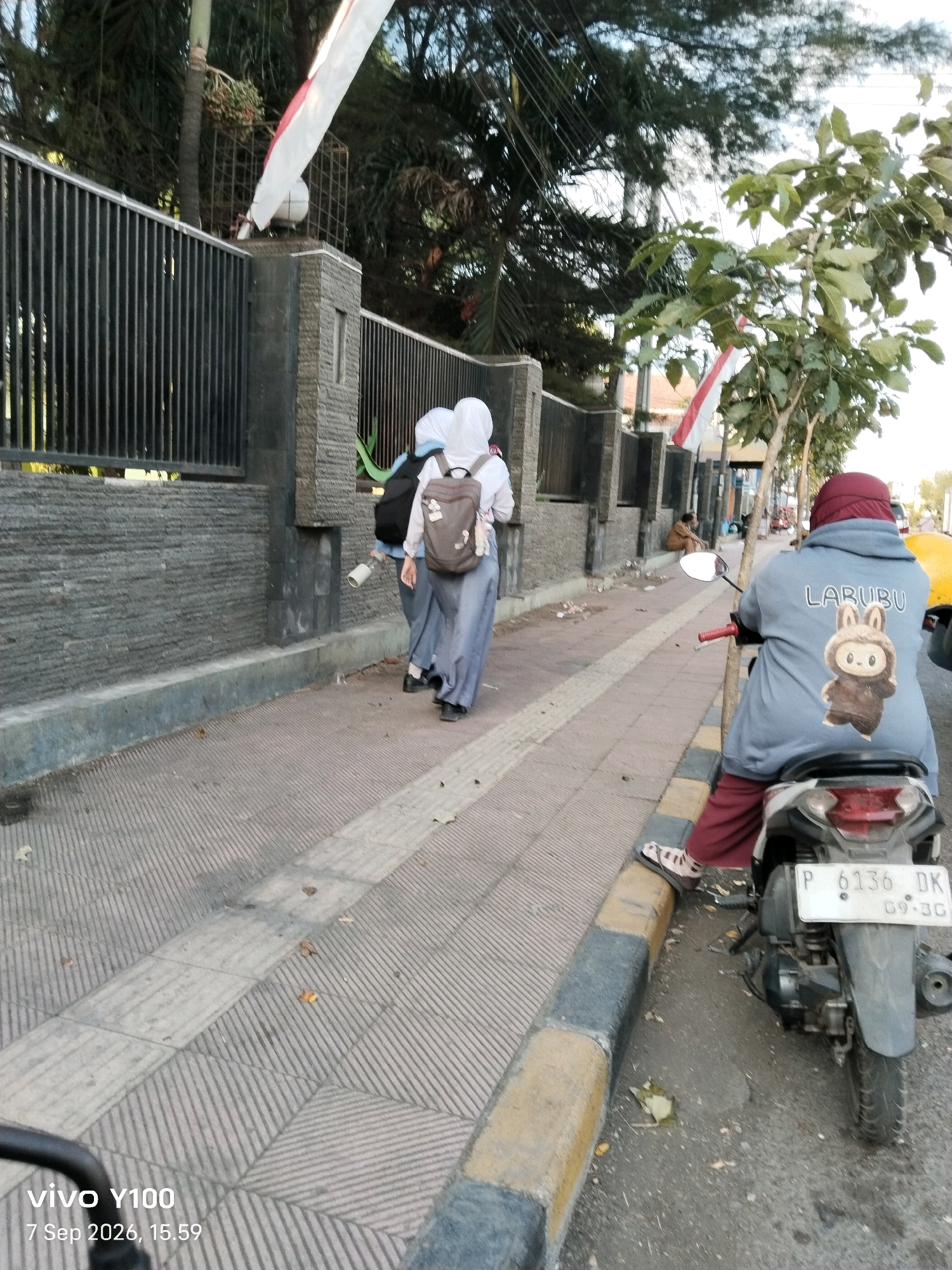Two young women in school uniforms and hijabs walk along a sidewalk next to a dark metal fence. One woman has a backpack. A person wearing a burgundy headscarf and a blue jacket with a cartoon bunny and the word "LARUBU" on the back sits on a scooter parked on the street next to the sidewalk. The scooter has a license plate that reads "P 6136 DK 09-30". Another person sits further down the street in the background. The scene appears to be a public area with trees and a building visible. The time stamp on the image indicates it was taken on September 7, 2026, at 15:59. The image includes a "vivo Y100" watermark. The location is identified as Situbondo, Indonesia.