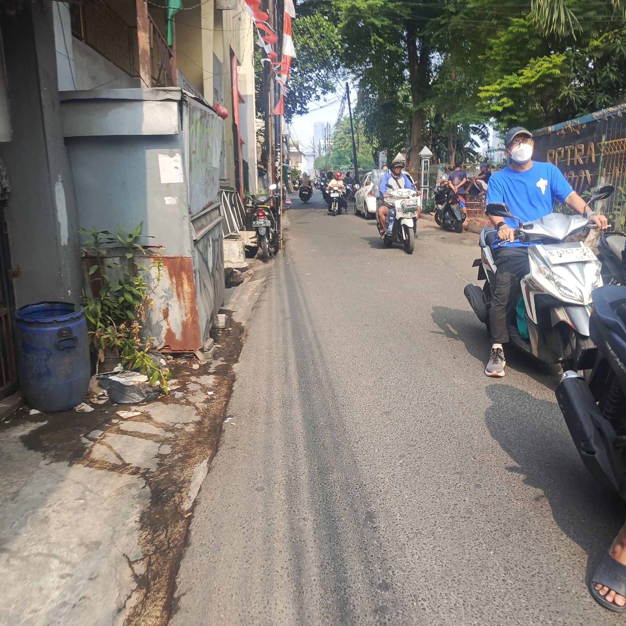 The image shows a street scene in South Jakarta, Indonesia, during daylight hours. A group of people are riding motorcycles along a paved road. One man in a blue t-shirt and a face mask is prominently featured, riding a white scooter. The street is lined with buildings and trees. There is a blue trash barrel and some debris on the side of the road. The overall impression is of a typical urban environment with moderate traffic.