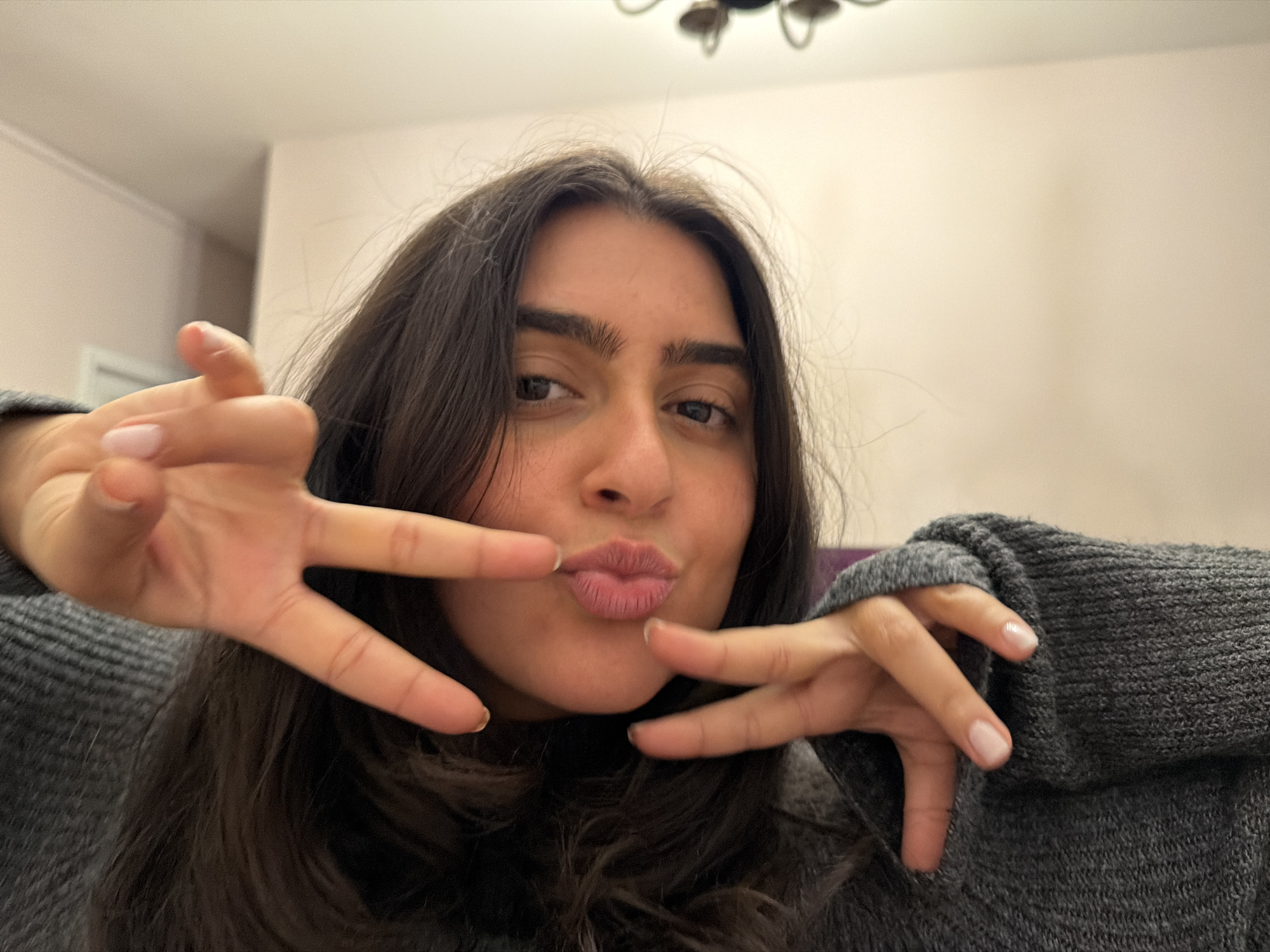 A woman with long, dark hair and light skin is holding her hands in front of her face, making a playful expression with pursed lips. She is wearing a gray sweater. The background is a light beige wall and a blurry chandelier is visible at the top. The location cannot be determined to be in New York City based on the image.