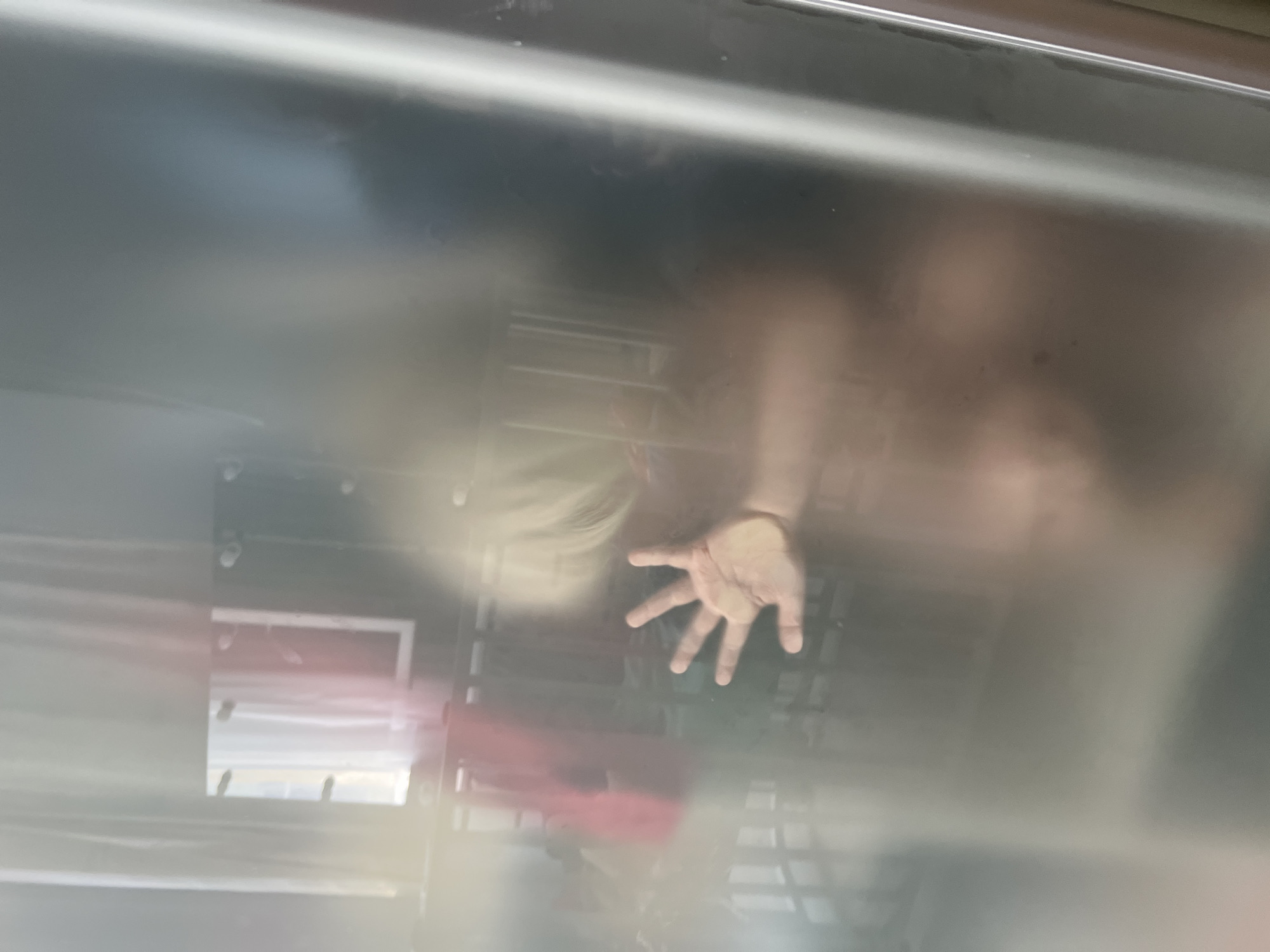 The image shows a blurry view through frosted glass or plastic, likely a shower door or enclosure. A child's hand is visible, splayed open, pressed against the surface. The child's arm and shoulder are also vaguely discernible, as is their blond hair. In the background, through the translucent barrier, a crib or bed with railings can be seen, along with some indistinct shapes that might be blankets or clothes. The scene appears to be indoors, in a domestic setting. There is no indication or information that this image is related to Sochi, Russia.