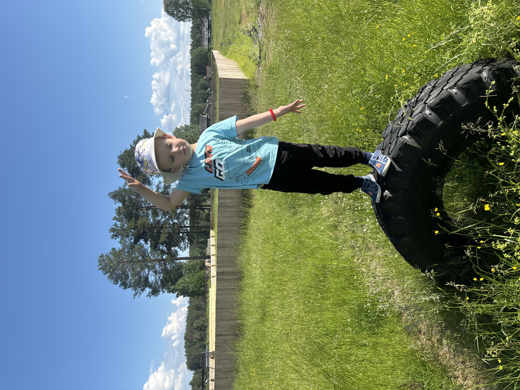 A young boy stands on a large tire in a grassy field under a bright blue sky. He is wearing a light-colored bucket hat with a pattern, a blue t-shirt with a graphic print, black pants, and blue sandals. His right arm is raised, with his index and middle fingers extended in a peace sign. The background features a wooden fence, trees, and some rural buildings. The image appears to be taken during the daytime in an outdoor setting. The location is in Kurgan, Russia.