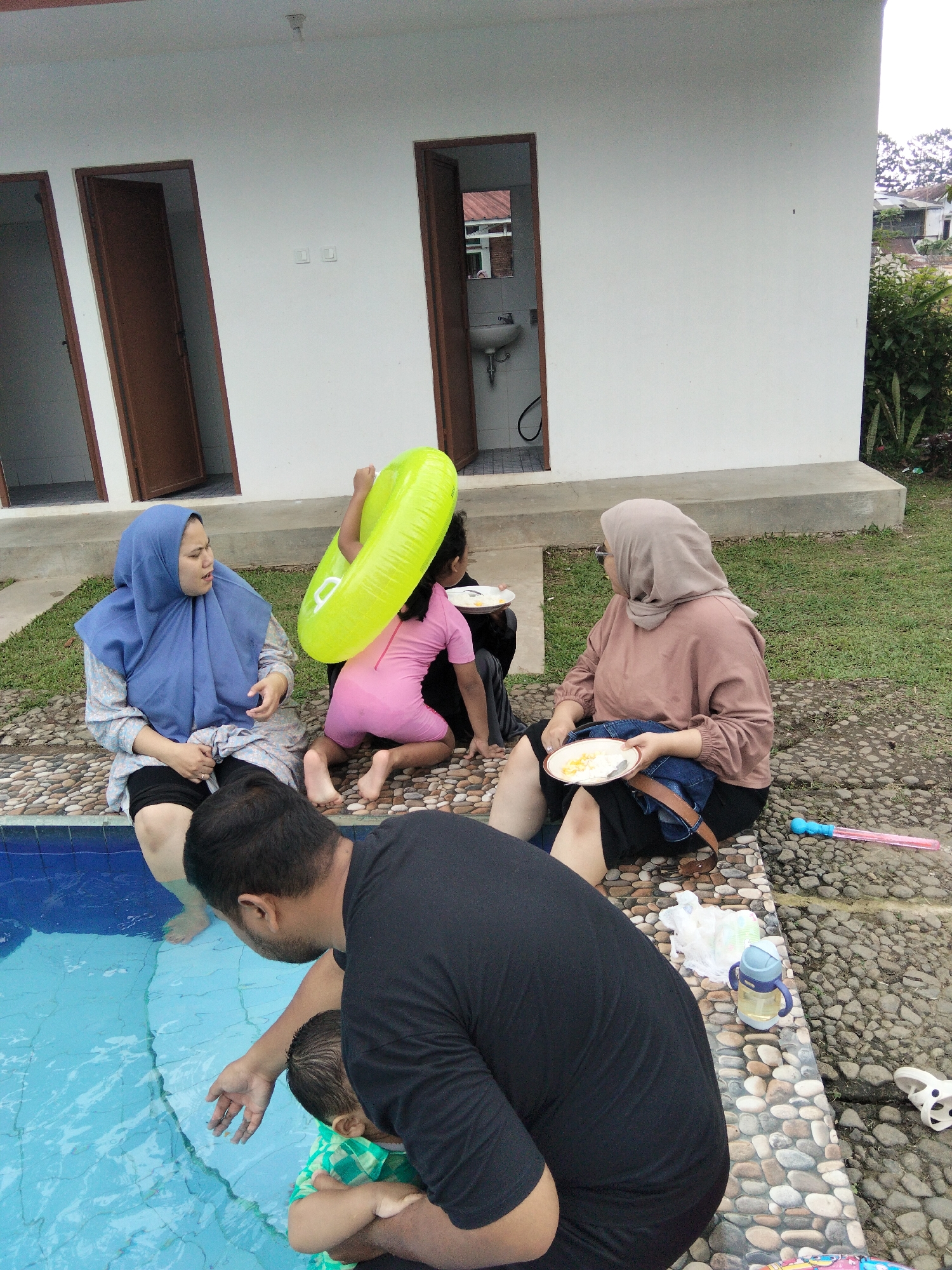 The image depicts an outdoor scene near a swimming pool in Cisarua, Indonesia. Five people, including two adults, two children, and one infant, are present. One adult woman wearing a blue hijab is seated on the edge of the pool, her legs bare. Beside her, a young child in a pink swimsuit is kneeling and holding a bright green inflatable ring. Another adult woman wearing a light brown hijab is seated on the ground further away, holding a plate of food. A man in a black t-shirt is partially visible in the foreground, holding an infant dressed in a green and white checkered shirt. The infant is reaching towards the pool water, and the man appears to be assisting the infant. The environment includes a swimming pool with clear blue water, a paved area surrounding the pool with decorative stones, and a building with multiple doors in the background. Lush green foliage is visible to the right of the building. A few objects are scattered on the ground, including a pink bubble wand and a blue and yellow water bottle. The overall atmosphere suggests a casual family gathering or outing.