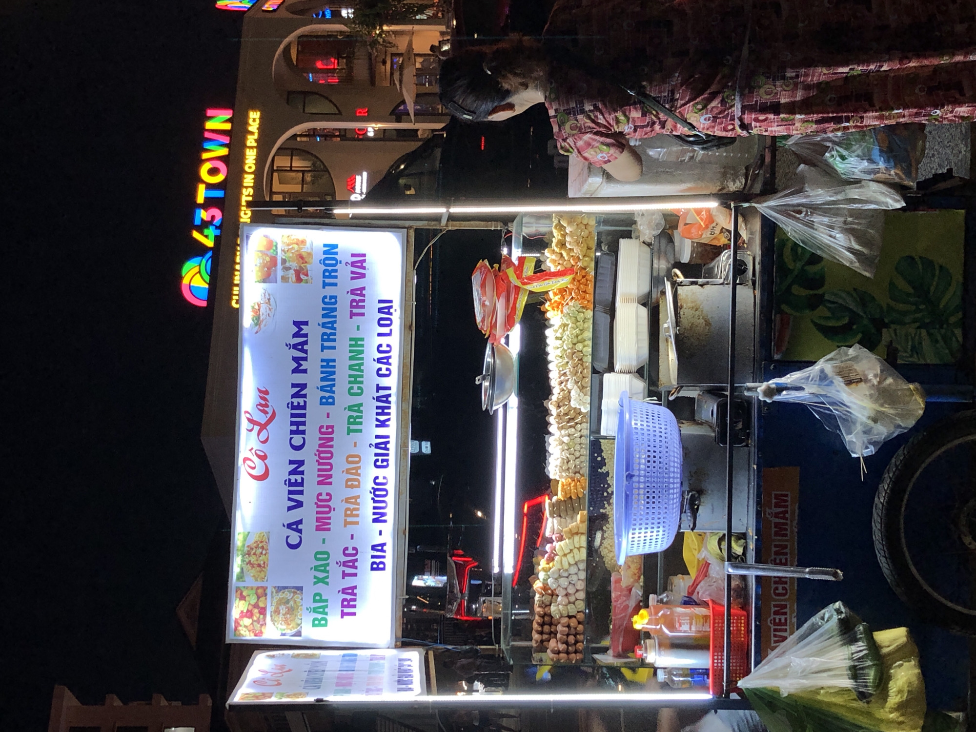 The image shows a night scene in Đà Nẵng, Vietnam, likely on a street with a commercial building in the background. In the foreground, a food cart is prominently displayed. The cart features a brightly lit sign with Vietnamese text and images of food. The text on the sign advertises "Cá Viên Chiên Mắm" (fried fish balls with fish sauce) and lists other items like "Bắp Xào" (corn stir-fry), "Mực Nướng" (grilled squid), "Bánh Tráng Trộn" (mixed rice paper), and various drinks including "Trà Tắc" (kumquat tea), "Trà Đào" (peach tea), and "Trà Vải" (lychee tea).
The cart itself is laden with food items. On display are skewers of what appear to be fried snacks, possibly fish balls and other processed meats, arranged in sections. There are also containers of pre-packaged food items and condiments. A blue plastic basket is placed on top of a metal container, and several stacks of white disposable food containers are visible.
A woman, seen from the back and side, is standing behind the food cart, appearing to interact with the items or customers. She is wearing a patterned dress. In the background, the building has a prominent neon sign that reads "43TOWN" with colorful lettering. Below this, another sign states "CULINARY LIGHTS IN ONE PLACE". The overall environment suggests a bustling street or market area at night.