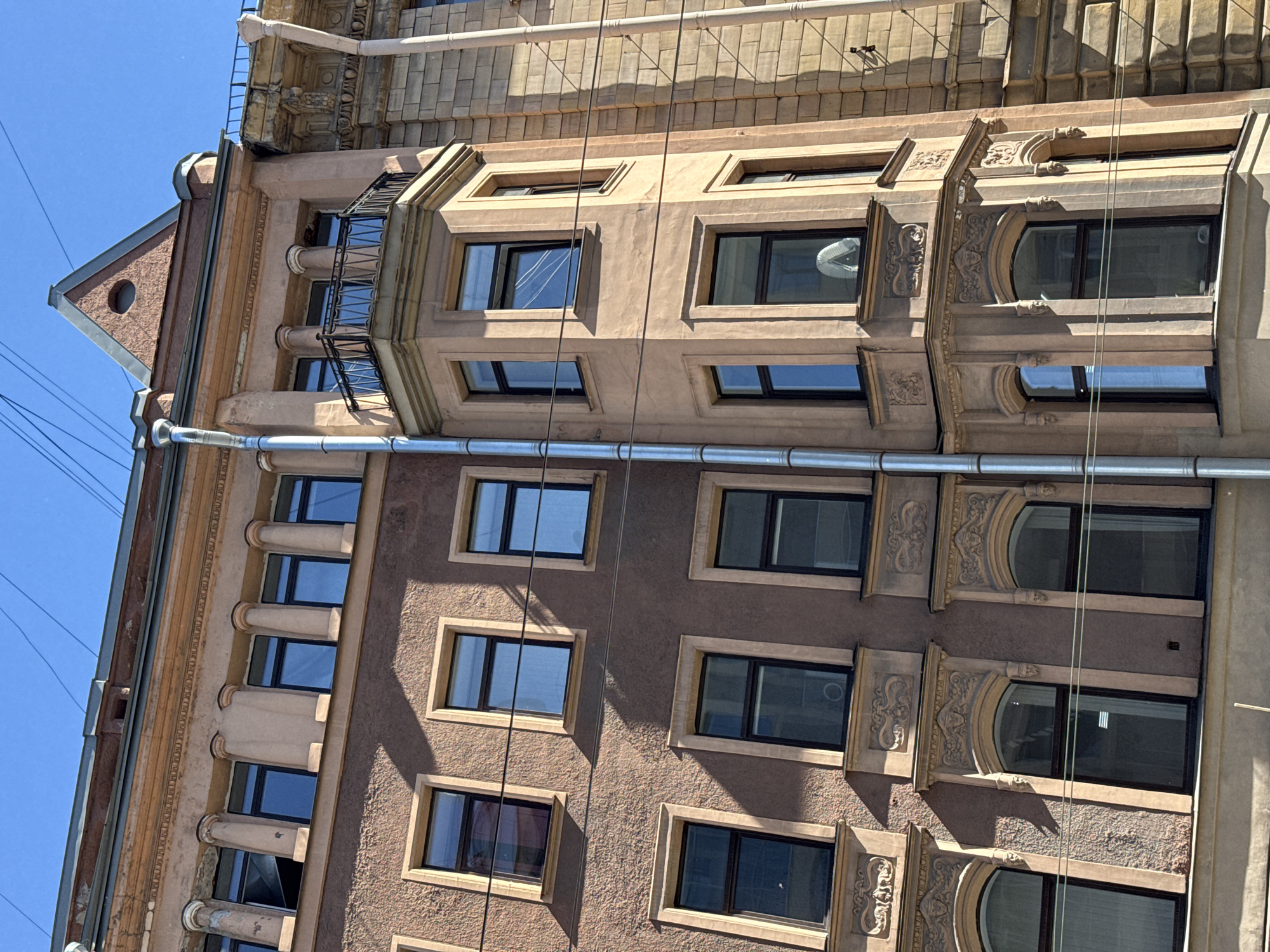 A low-angle, eye-level exterior shot captures the facade of a multi-story building with an ornate architectural style under a clear blue sky. The building features a light brown, stucco-like texture on its walls, contrasted with darker brown window frames and lighter beige decorative trim around the windows and along the roofline.
The building's facade is punctuated by numerous rectangular windows with dark frames. Some windows are a solid dark color, while others reflect the blue sky, indicating they are glass. The second floor from the top displays a series of paired pillars above the windows, separated by sections of the stuccoed wall. The topmost floor has a section with a pointed, triangular roof and a small circular window.
A prominent feature is a bay window area on the right side of the building, which juts out from the main facade. This bay window has multiple panes and is adorned with decorative carvings above some of the windows. A black metal railing outlines a small balcony that sits atop this bay window section.
A large, corrugated metal pipe runs vertically down the center of the building, seemingly a drainpipe. Above this pipe, the building's roofline is visible, with a slightly damaged or unfinished section at the very top. Electrical wires are strung across the sky and in front of the building, crossing over the windows.
The lighting suggests it is daytime, with shadows cast by the architectural elements and the windows onto the building's surface. The overall impression is one of old-world architecture, possibly in a European city, with a somewhat weathered but still grand appearance. There are no people visible in the frame.