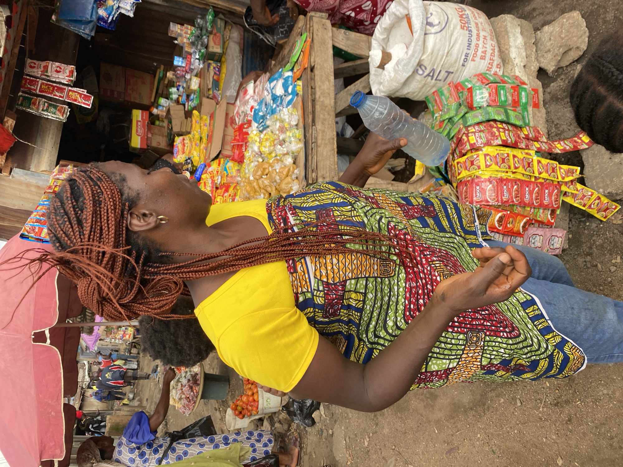 A woman with braided brown hair and a yellow shirt stands in a bustling outdoor market in Bukuru, Nigeria. She wears a colorful, patterned dress and blue jeans, and holds a clear plastic water bottle. In front of her are rows of small, individually packaged food items, primarily in red, yellow, and green wrappers. To her right, a large white sack labeled "SALT" sits on a stone. The background reveals more market stalls with various goods, including produce and packaged food, and other people browsing and working. The ground is dirt and debris, indicative of an active marketplace.