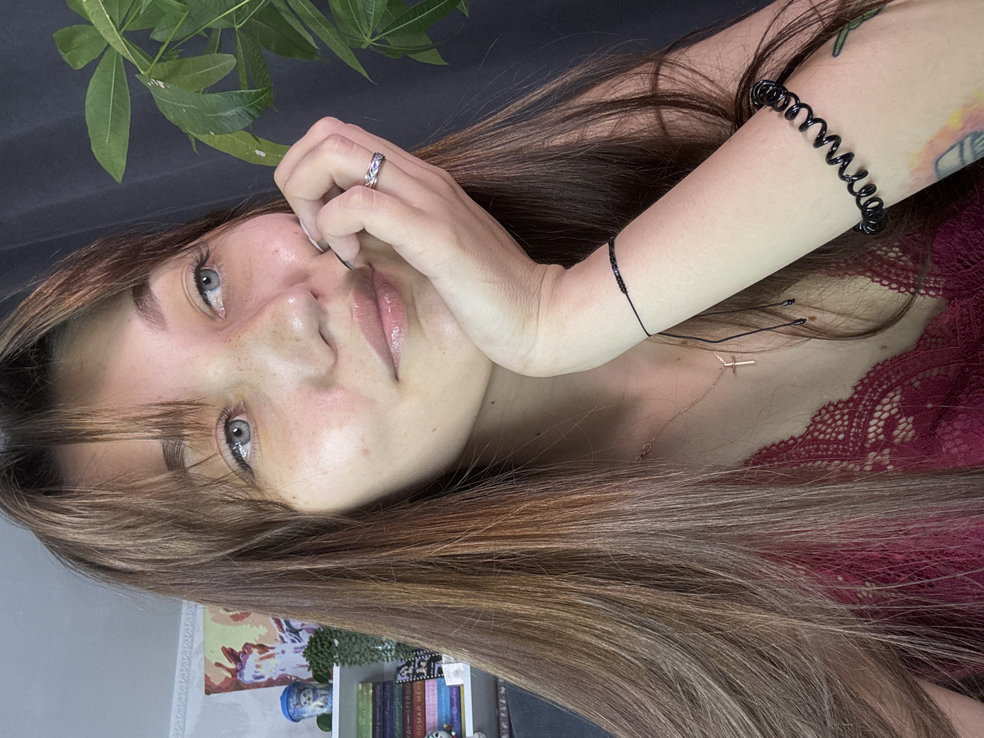 A young woman with long, brown hair is lying down, with her head tilted to the side. She is looking directly at the camera with a soft expression. Her hand is resting on her face, and she is wearing a ring on her finger. A black, spiral bracelet is on her arm. She is also wearing a pink lace top. A plant with green leaves is visible in the upper left corner of the image. In the background, there is a bookshelf filled with books, and some artwork on the wall. The overall lighting suggests it is daytime. The image appears to be taken indoors. There is no visible text in the image.