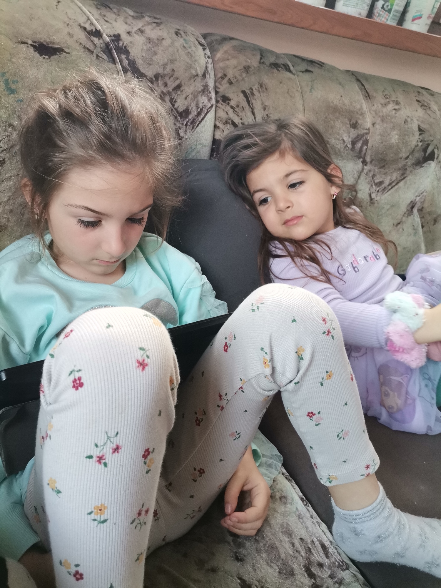 Two young girls are sitting on a couch. The girl on the left is wearing a light blue sweater and floral leggings and is focused on a black tablet. The girl on the right is wearing a lavender sweater and floral leggings with a patterned blanket. The couch is patterned and the background is indistinct. The location is Unknown, Unknown.