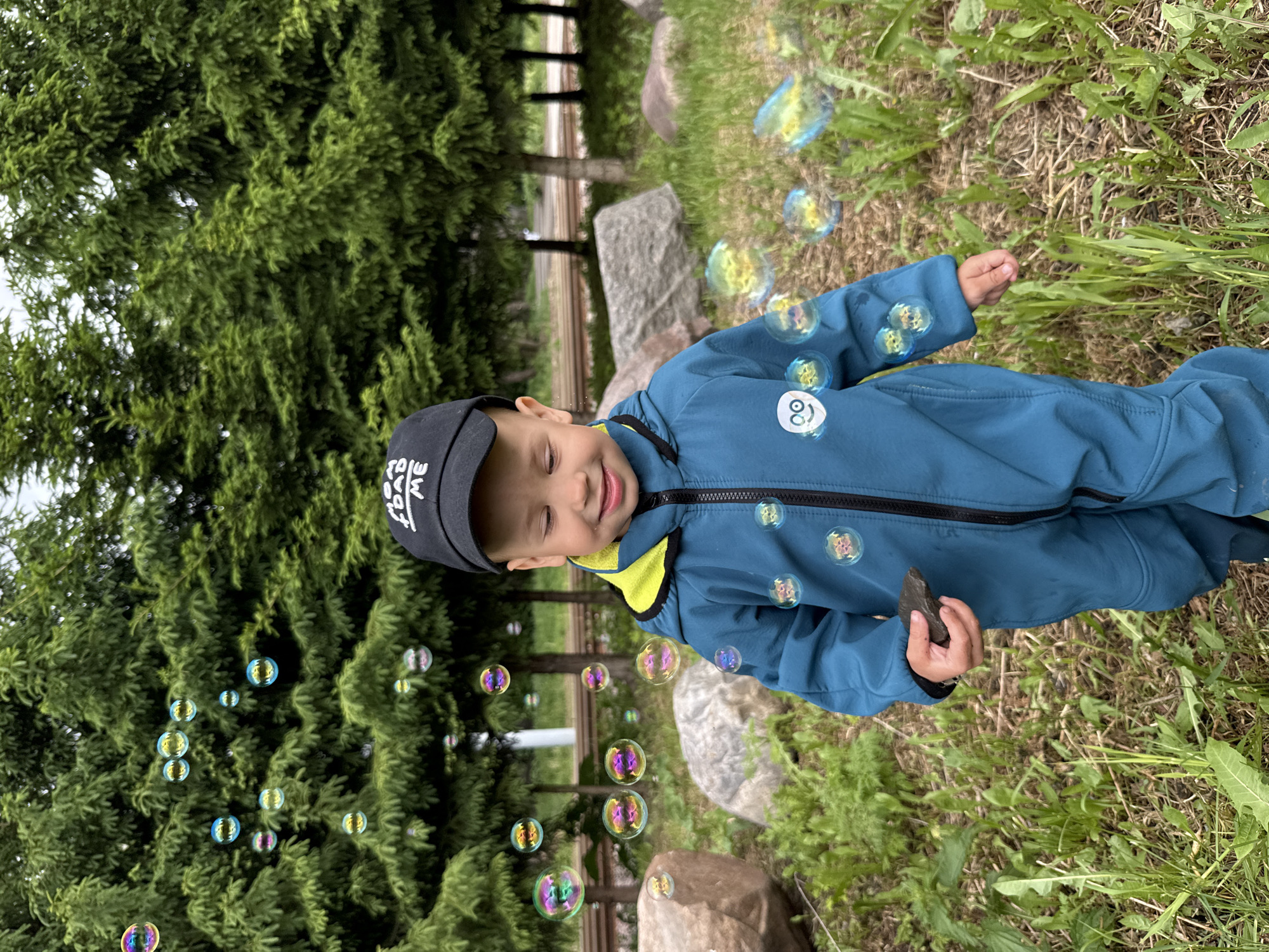 A young child wearing a black baseball cap and a blue jumpsuit stands in a grassy area with trees and large rocks. The child is holding a small, dark-colored rock in their right hand and is surrounded by numerous floating soap bubbles. The scene is outdoors, with the background consisting of trees, grass, and some fencing. The photograph appears to have been taken in Rusanovo, Russia.
