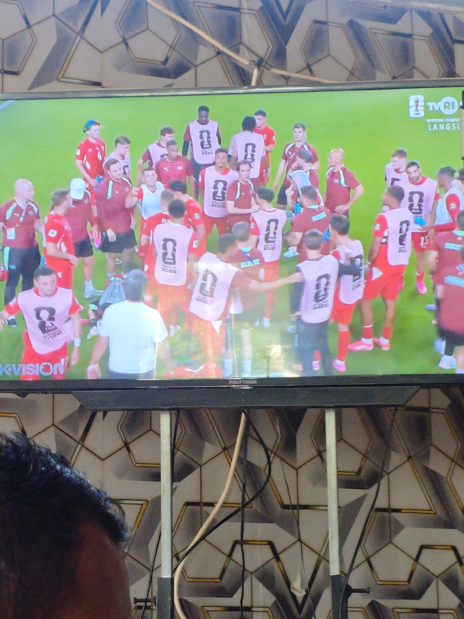 The image shows a television screen displaying a group of male soccer players on a green field, possibly a stadium. The players are wearing red and white uniforms, with some wearing pink bibs over their jerseys. One player in the foreground is wearing a white t-shirt. A logo with "TV RI" is visible in the top right corner of the screen, suggesting the broadcast is from Indonesia. The television is placed on a black stand, and behind it, a wall with a geometric pattern is visible. A person's head and shoulder are in the foreground of the image, obscuring the bottom part of the television screen and the stand. The overall scene appears to be a viewing of a sports event, likely in a home or public space in Parangsembilan, Indonesia, given the "TV RI" logo which is the public television broadcaster of Indonesia.