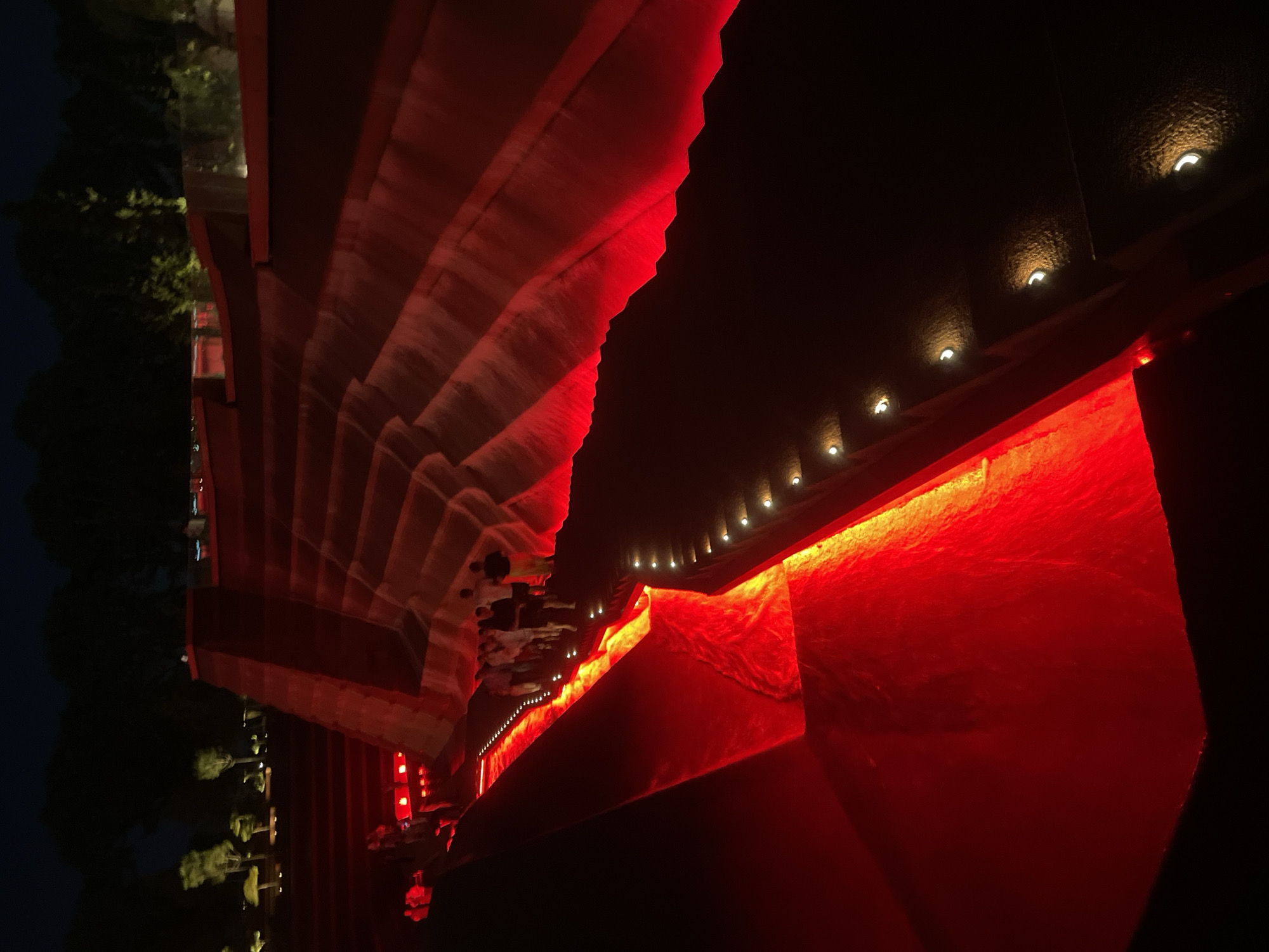 The image depicts a large, multi-tiered outdoor structure illuminated by vibrant red lights at night. The architecture features angled, layered surfaces that resemble steps or terraces, with a central channel of water flowing or cascading down. Small, bright lights are embedded along the edges of the steps, providing additional illumination. A group of people, appearing to be tourists, are visible walking along the pathways and stairs, some pausing to observe the scene. In the background, dark silhouettes of trees suggest an outdoor park or garden setting, characteristic of the city of Krasnodar, Russia, particularly its park areas. The overall impression is of a modern, artistic light installation within a public space.