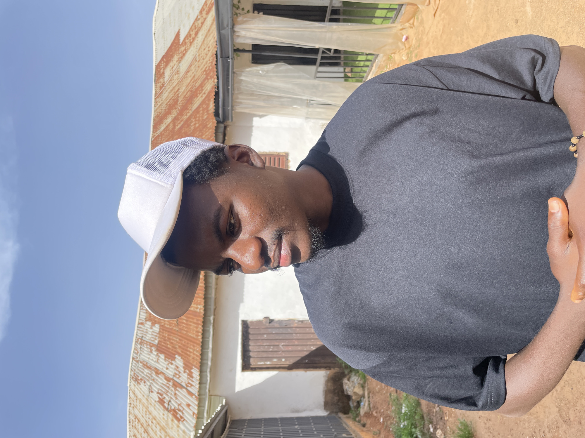 A close-up, tilted shot features a dark-skinned man wearing a white baseball cap and a dark gray t-shirt. He is looking downwards with a serious expression. A portion of a house is visible in the background. The house has a corrugated metal roof, white walls, and a window covered with what appears to be a thin, transparent plastic or fabric. In the background there is also some ground and vegetation, possibly of a garden. From the angle, it looks like a street scene in Barakin Akawo, Nigeria.