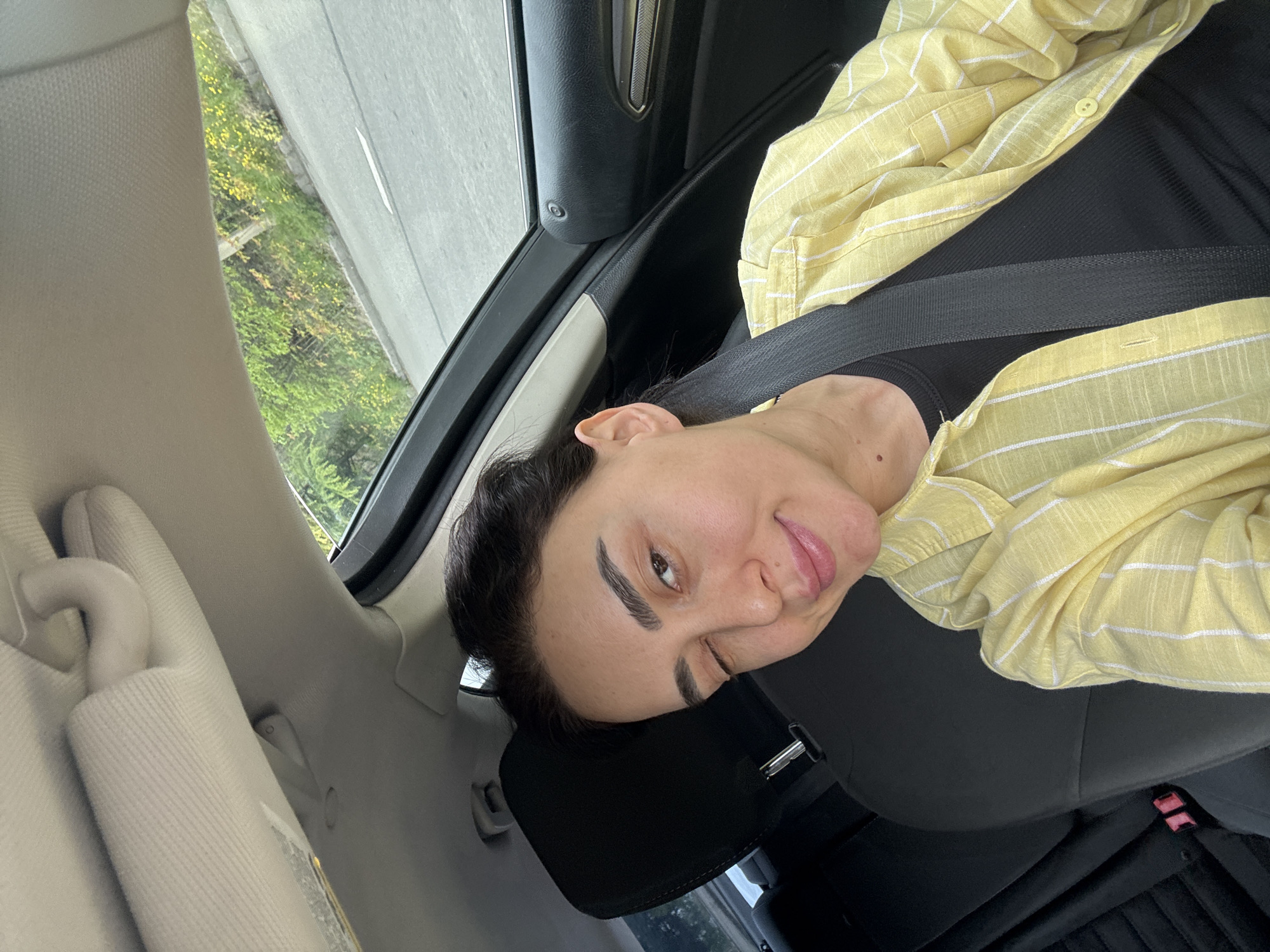 The image, initially rotated 90 degrees clockwise, captures a close-up of a woman in the passenger seat of a car during the daytime.
The primary subject is a woman with dark hair, dark eyebrows, and a light complexion. She is wearing a light yellow button-up shirt with thin white stripes over a black top, secured by a black seatbelt across her chest and lap. Her head rests on a black headrest, and she is looking directly at the camera with a relaxed expression, featuring a slight smile and one eye appearing to wink or be partially closed, suggesting a playful or casual mood.
The setting is the interior of a car. Visible car elements include a light beige/tan sun visor in the foreground, black car seats, and parts of a black door panel and window frame. Through the car window, a bright outdoor scene is visible, featuring lush green foliage with hints of small yellow flowers, suggesting a natural environment alongside a grey road or pavement.
The activity taking place is the woman posing for a photograph, likely a selfie, while traveling as a passenger in the car. The time of day is clearly daytime, indicated by the natural light illuminating both the car interior and the external scenery. There are no specific visual cues within the image itself to confirm the provided location context of Tbilisi, Georgia. No readable text is visible in the frame.