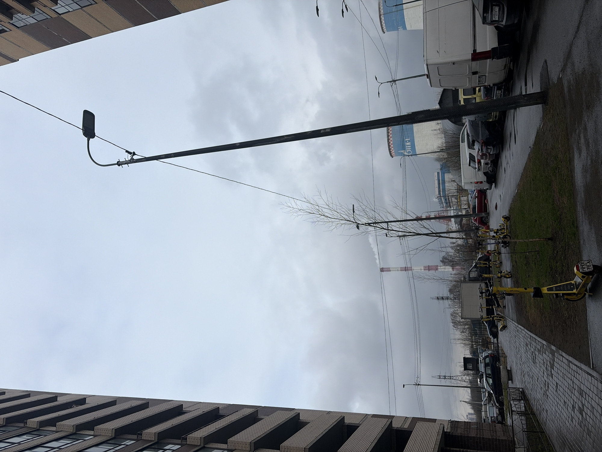 The image depicts a street scene in Saint Petersburg, Russia, on a cloudy day. The view is oriented upwards, capturing a streetlamp and power lines against the grey sky. Two buildings, one with a distinctive textured facade, frame the top and bottom of the image. Along the right edge of the frame, a sidewalk is visible, with a line of parked electric scooters and cars. In the background, industrial chimneys rise above the horizon, suggesting an urban industrial zone. The overall atmosphere is overcast and somewhat bleak.