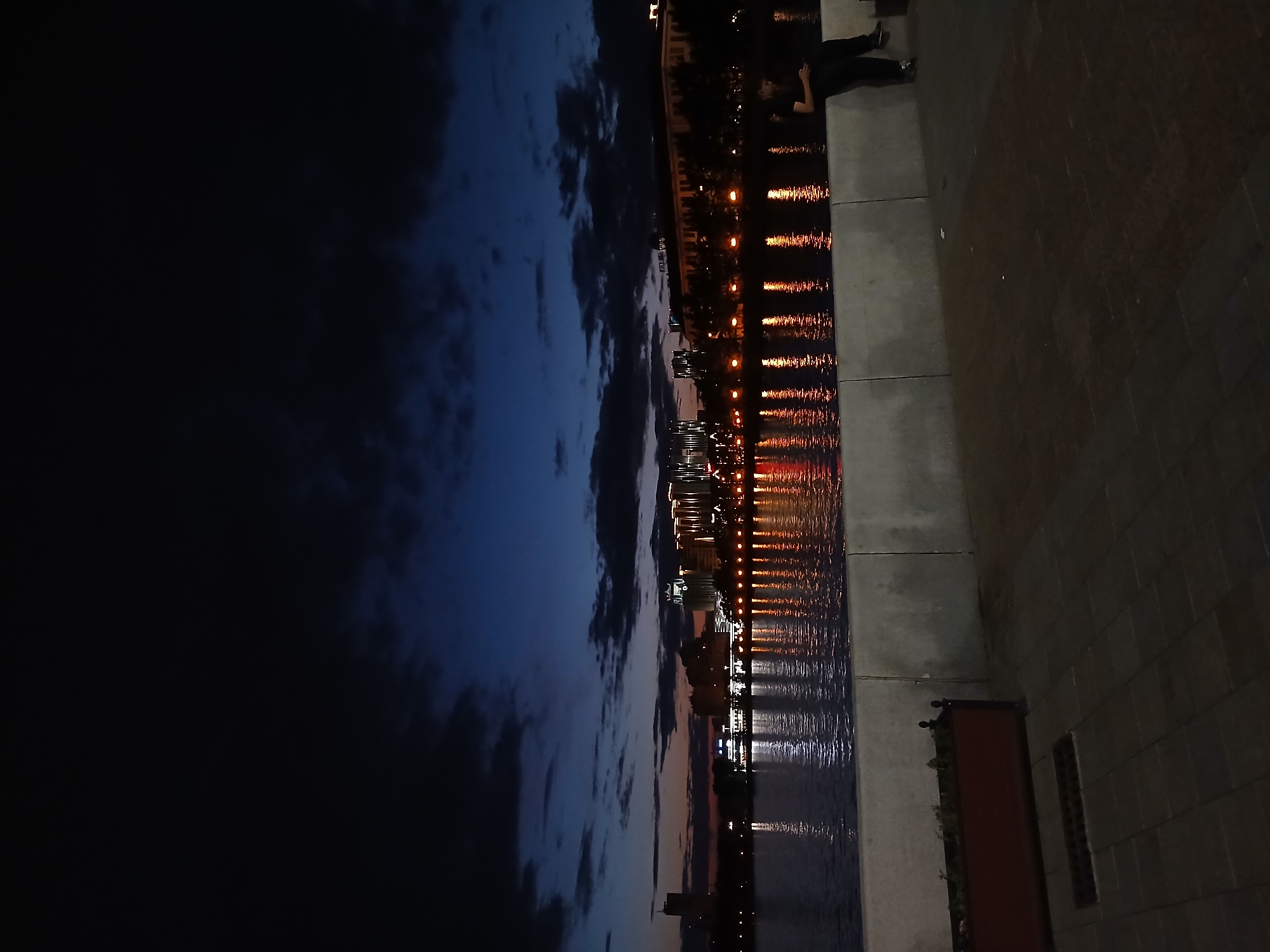 The image captures a twilight scene along the Iset River in Yekaterinburg, Russia. On the opposite bank, a brightly lit cityscape with various buildings and structures is visible, their reflections shimmering on the water's surface. In the foreground, a person is seated on a concrete embankment, looking down at a mobile phone. The area is paved with dark tiles, and a planter box with sparse greenery is positioned to the left. The sky above is a deep blue, transitioning to darker shades, with clouds adding texture to the horizon.