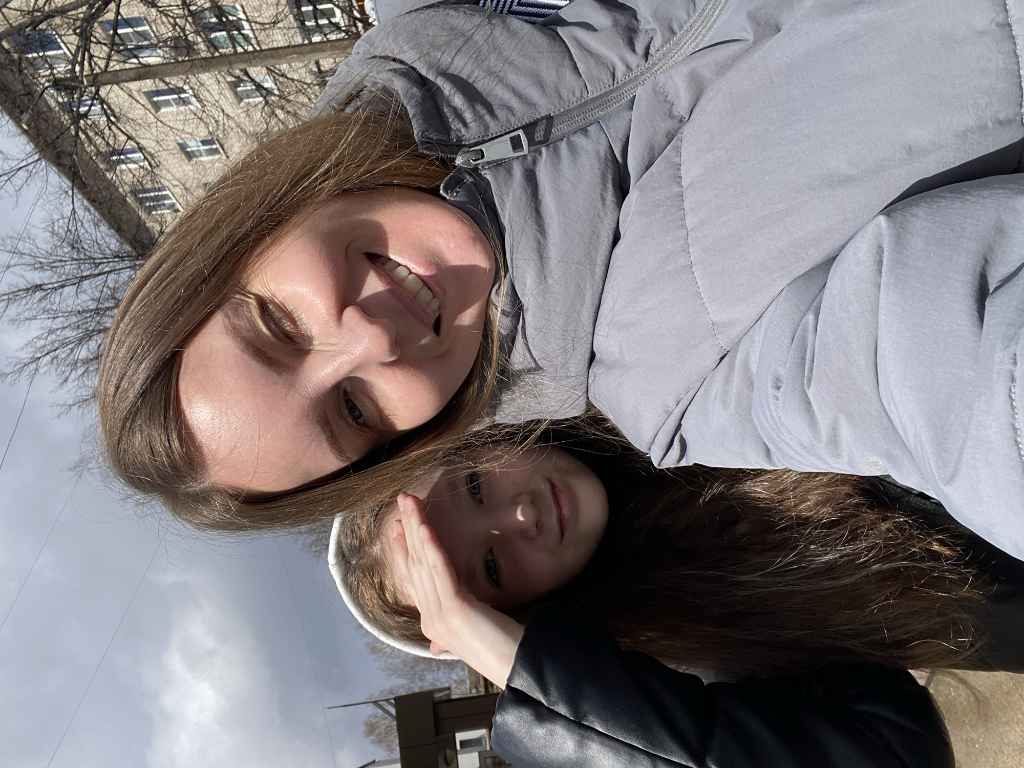 The image depicts a selfie of two individuals. The first is an adult woman with fair skin, light brown hair, and wearing a light gray puffer jacket. She is smiling. Below her is a younger girl with fair skin and long brown hair. She is wearing a white headband and a black leather jacket. Her hand is raised to shield her eyes. They are outdoors on a sunny day. In the background is a multi-story building with bare trees. The city is Chaikovsky, Russia.