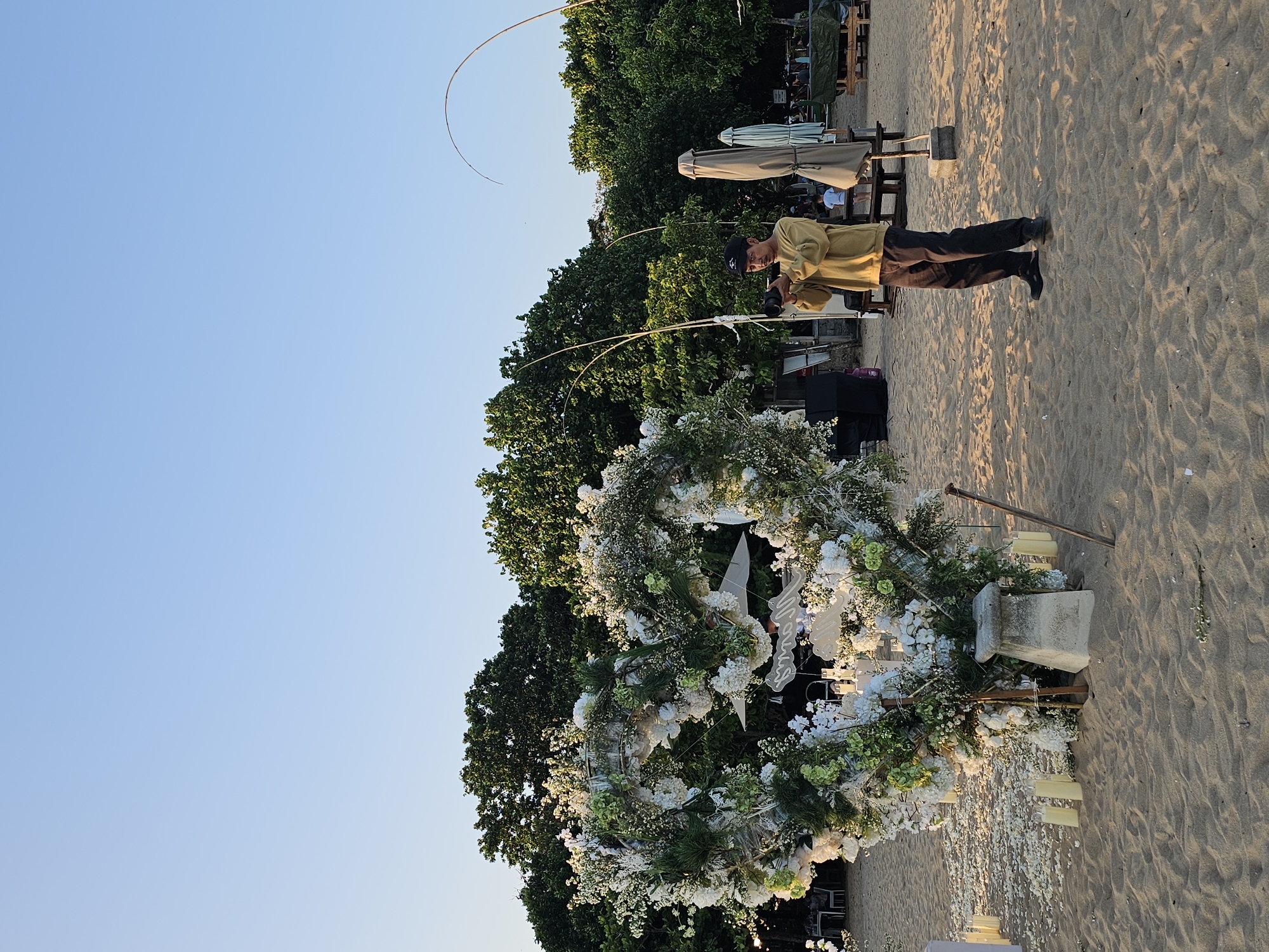 A man is photographing a wedding arch on a sandy beach. The arch is round and adorned with white flowers and greenery. The man is wearing a yellow shirt and dark pants. Behind the arch are trees. The scene is set on a beach in Denpasar, Indonesia. There are parasols and other beach furniture visible in the background.